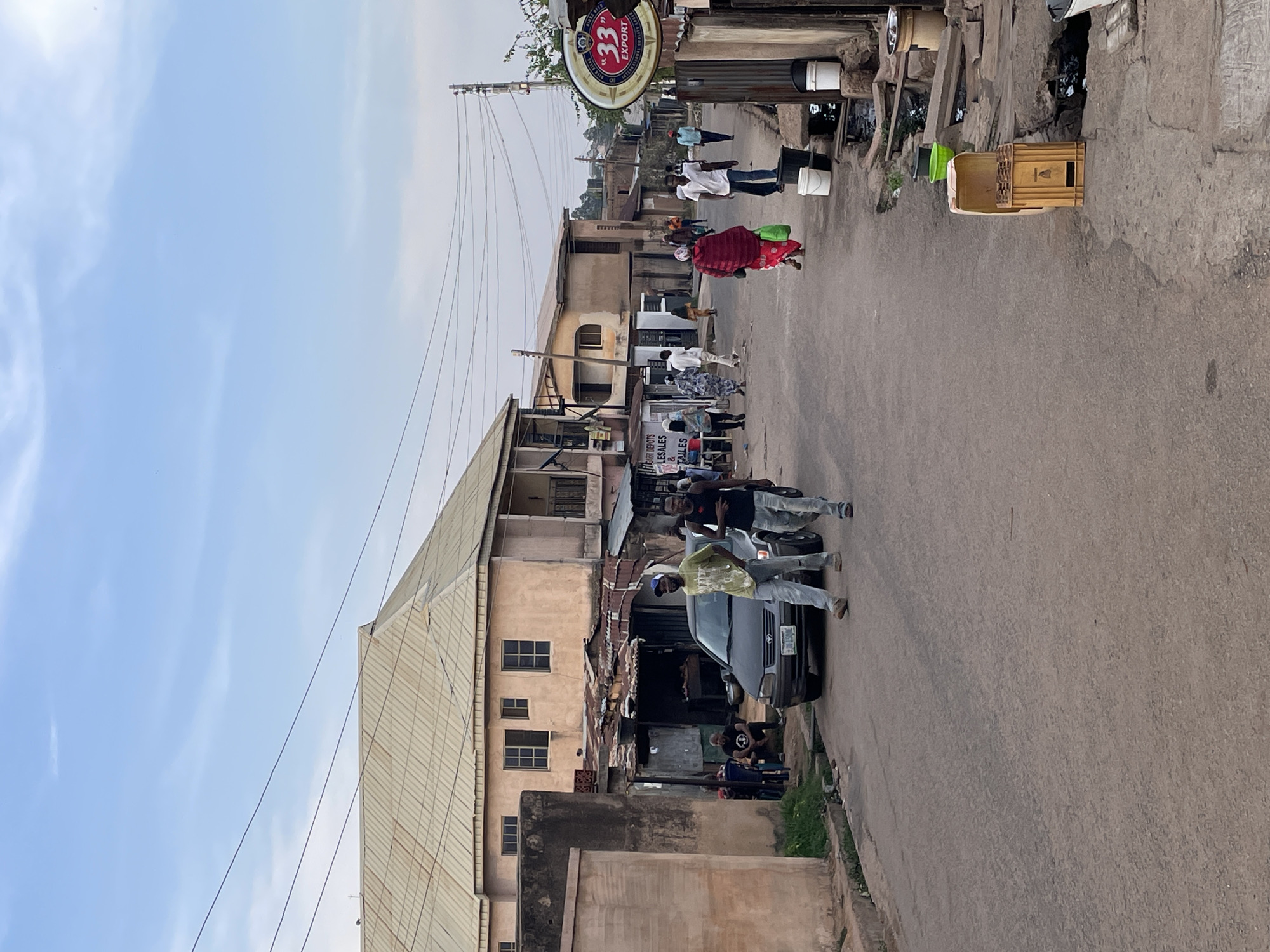 This rotated image captures a lively street scene in Jos, Nigeria, under a clear, bright blue sky with sparse clouds, indicating daytime. The paved street is flanked by buildings of varying heights and construction styles, many appearing weathered with some featuring corrugated metal roofs.
Multiple people are present, engaged in daily activities. On the right side of the street, several individuals are walking, including one person in a red and dark patterned outfit carrying a green bag. Nearby, two men stand beneath a prominent circular sign that reads "33 EXPORT," with smaller text identifying "NIGERIAN BREWERIES PLC" and "LAGOS NIGERIA." Utility poles and overhead power lines are visible along the street.
On the left, a dark-colored car is either parked or moving slowly. Several men are gathered around it, including one in a light yellow-green t-shirt and blue cap, and another in a black t-shirt. Another individual is seated on the ground further back. Along the roadside, various objects like white buckets and a wooden crate with a green lid are present. A partially visible sign in the mid-ground appears to read "DEPOT BOTS LESALES & TAILLES," suggesting commercial establishments. The overall atmosphere depicts an ordinary, bustling day in an urban or semi-urban setting.