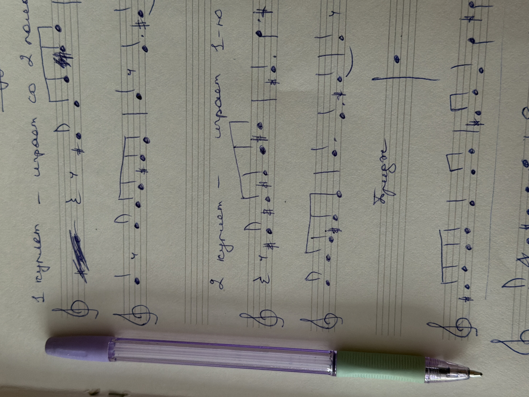 The image shows a handwritten musical score on lined paper, with a lilac-and-green pen lying horizontally across the bottom of the page. The music is written in blue ink and appears to be a treble clef melody. Markings in Russian indicate that the music is intended for "1 куплет" (1st verse) and "2 куплет" (2nd verse), to be played "со 2 голоса" (with the 2nd voice). Further annotations such as "играет" (plays), "I-й играет" (1st plays) and "легче" (easier) are also visible. The environment appears to be indoors, with natural light illuminating the page. There is no location information available in the image.
