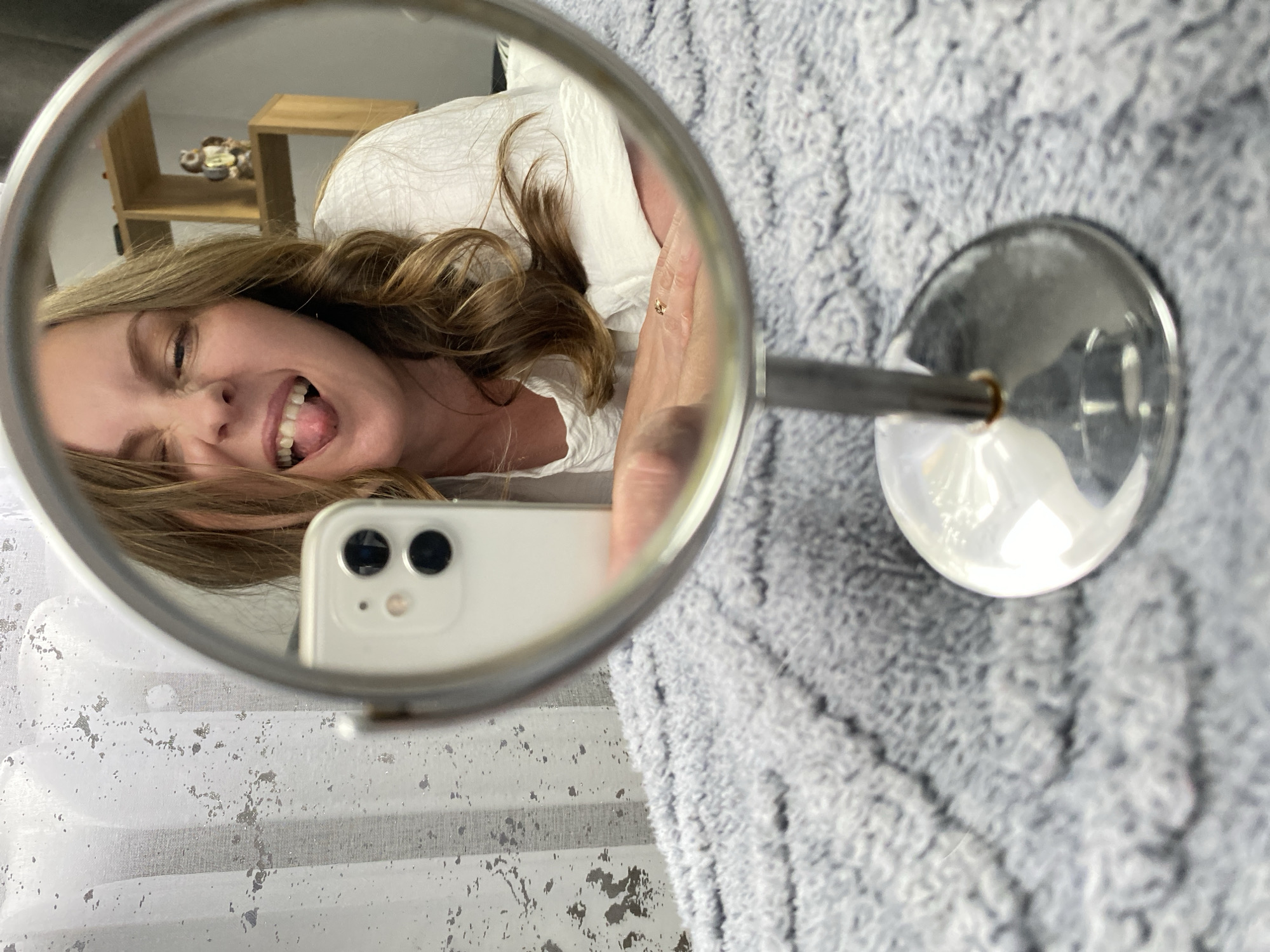 A woman is taking a selfie in a handheld mirror. The reflection shows the woman's face, shoulder, the phone and hand holding the phone. The phone is white and has two cameras. The woman is sticking her tongue out. There is a partially visible wooden shelf in the background and a gray textured fabric surface. The location is not discernible from the image.