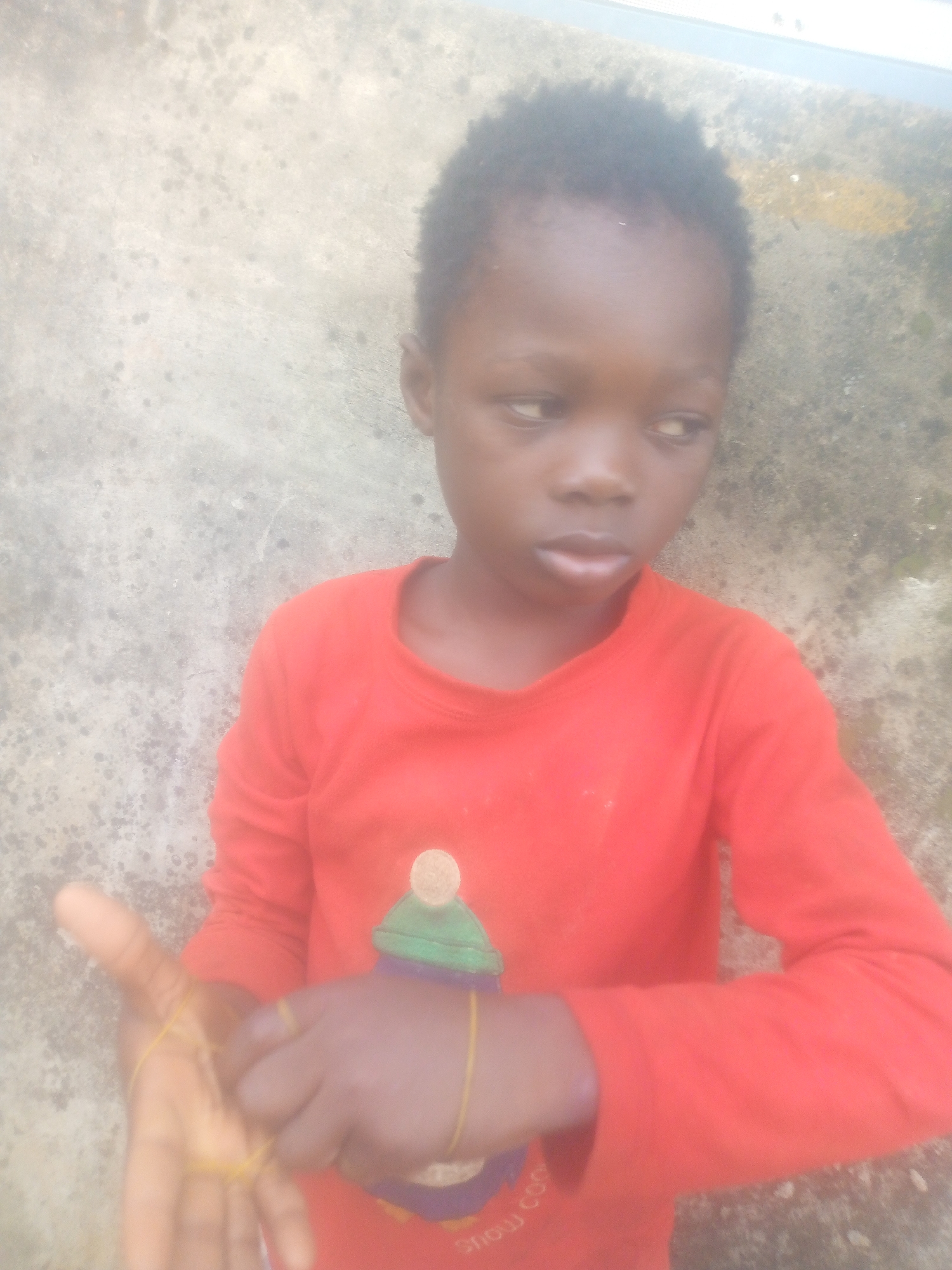 A young child, likely between 5 and 10 years old, with dark skin and short, dark, curly hair, is depicted in a full-frame portrait. The child's gaze is directed slightly off-camera to their right, with a neutral expression. They are wearing a bright red long-sleeved shirt featuring a graphic design on the front. The graphic appears to be a stylized green winter hat or cap with a lighter, possibly yellow or beige, pompom. Below the graphic, some blurry text is visible, with parts of what might be "snow" or "show coo" discernible.
The child's hands are prominent, with several yellow rubber bands stretched across their fingers, suggesting an activity or game involving the bands.
The background is a textured wall, appearing to be concrete or stucco, mottled with shades of grey and light beige. It shows some irregular darker spots or stains. The lighting is soft and diffused, indicating daytime conditions, possibly overcast or in a shaded outdoor setting. There are no specific cues to identify the exact location.