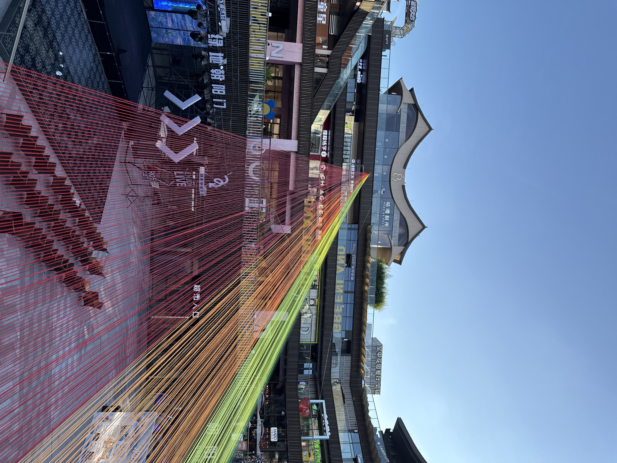 This is an outdoor, elevated shot of the "Rose Garden" shopping mall in Yiwu, China, on a clear day. The foreground is dominated by a large, colorful installation of red, orange, and green strings that converge towards the top of the frame, creating a triangular or conical shape. Below the strings, on the ground level, are rows of dark red chairs arranged in a stepped pattern, facing a stage area. The stage is set up with lighting equipment and a large screen displaying abstract blue and white imagery. To the left of the stage, there are large white arrow-like graphics.
The midground features the multi-story architecture of the Rose Garden mall. The building has a modern design with layered balconies, glass facades, and distinctive, curved, pagoda-style roofs. Various signs with Chinese text are visible on the building's exterior, advertising shops and services. People are seen on the balconies and walkways of the mall, observing the scene below.
The background consists of a clear, bright blue sky. The overall impression is of a lively, contemporary urban space, likely a public area within the mall, set up for an event or performance.