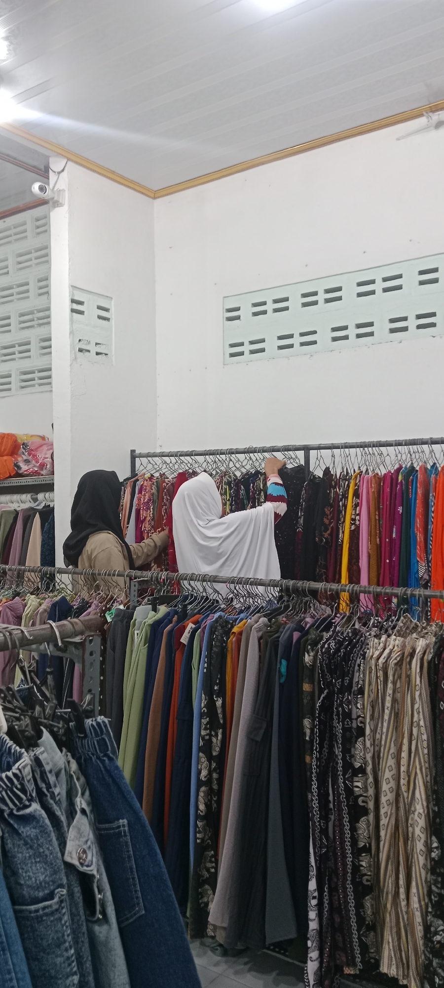 In a retail establishment in Rengel, Indonesia, two individuals are observed browsing through clothing racks. One person, wearing a black hijab and a tan garment, is positioned on the left side, examining items on a lower rack. To their right, a second person, entirely covered in white Islamic attire, is reaching for an item on a higher rack. The environment is characterized by multiple clothing racks filled with a variety of garments, predominantly dresses and skirts, displaying diverse patterns and colors. The store appears to have white walls and a white paneled ceiling with fluorescent lighting. Vents are visible on the upper portion of the walls. In the foreground, denim jeans are displayed on a separate rack. A security camera is mounted on a pillar near the ceiling in the upper left corner of the image.