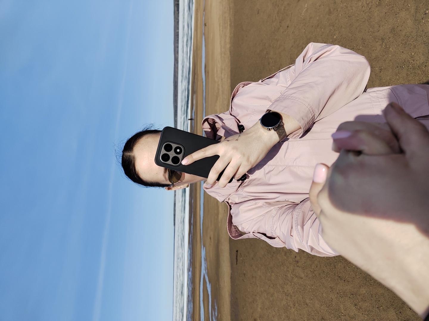 A woman is holding a black phone in her left hand, with a watch visible on the wrist of the same arm. The background shows a beach scene with sand and water, under a blue sky. The photo was taken in Chaikovsky, Russia. Someone is holding her hand. She is wearing a light pink jacket.