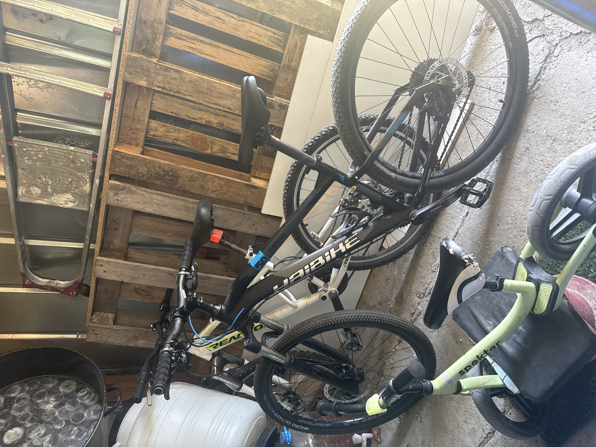The image shows a cluttered storage space in Elhovo, Bulgaria. Two bicycles, a black Haibike and a white Reaction, are leaning against a rough, light-colored wall. Nearby is a child's "spekter" tricycle. To the left, a metal ladder and a wooden pallet are leaning against another wall. In the lower left corner, there is a large metal pot filled with circular objects. A white plastic barrel sits beside it. The floor appears to be concrete. There are no people visible.
