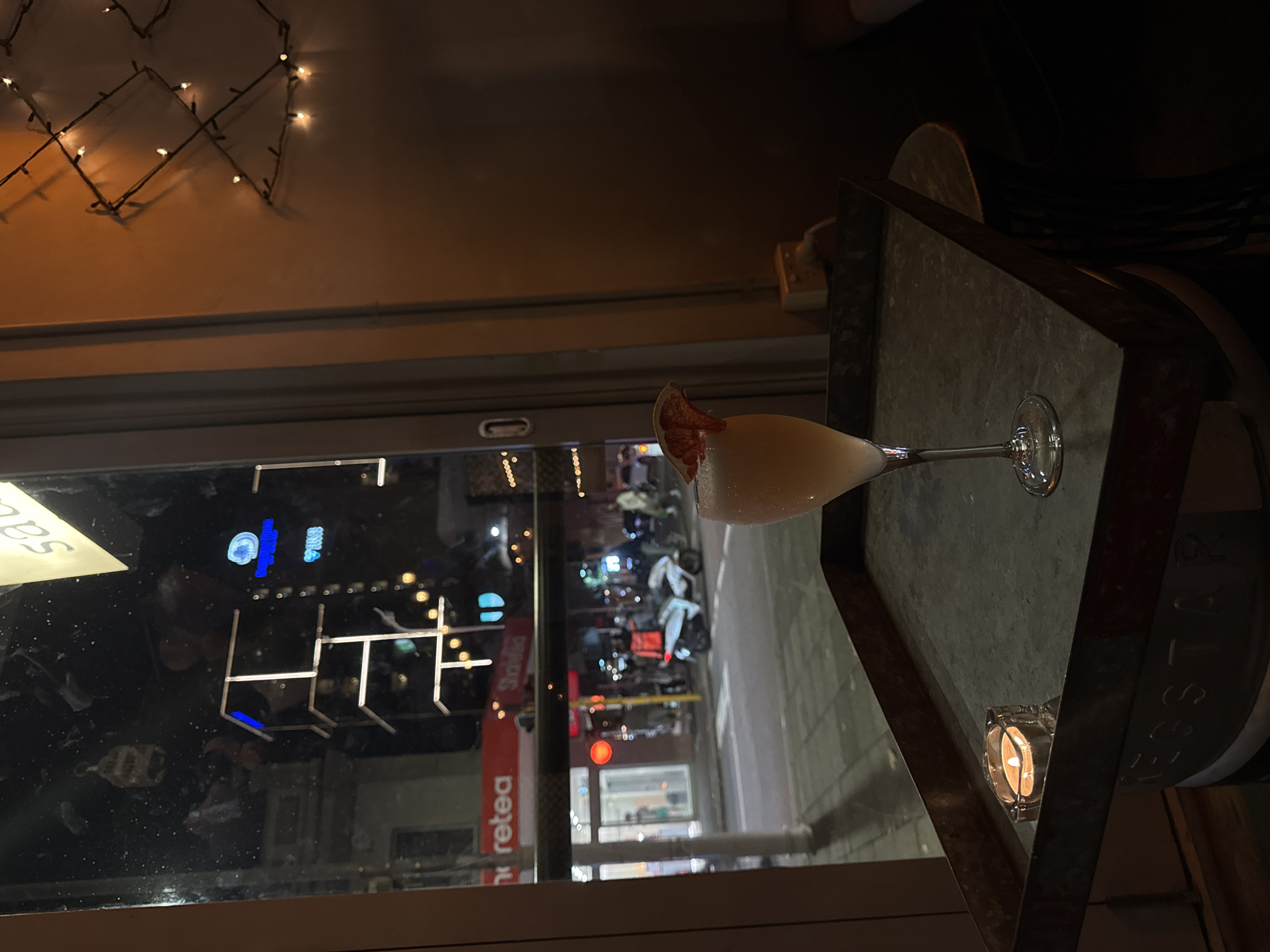 A cocktail glass sits atop a table indoors, near a window offering a view of a Northbridge street at night. The outdoor scene, with its lights and signs, indicates an urban environment. Reflected in the window is the interior, featuring fairy lights. The table also has a candle on it.