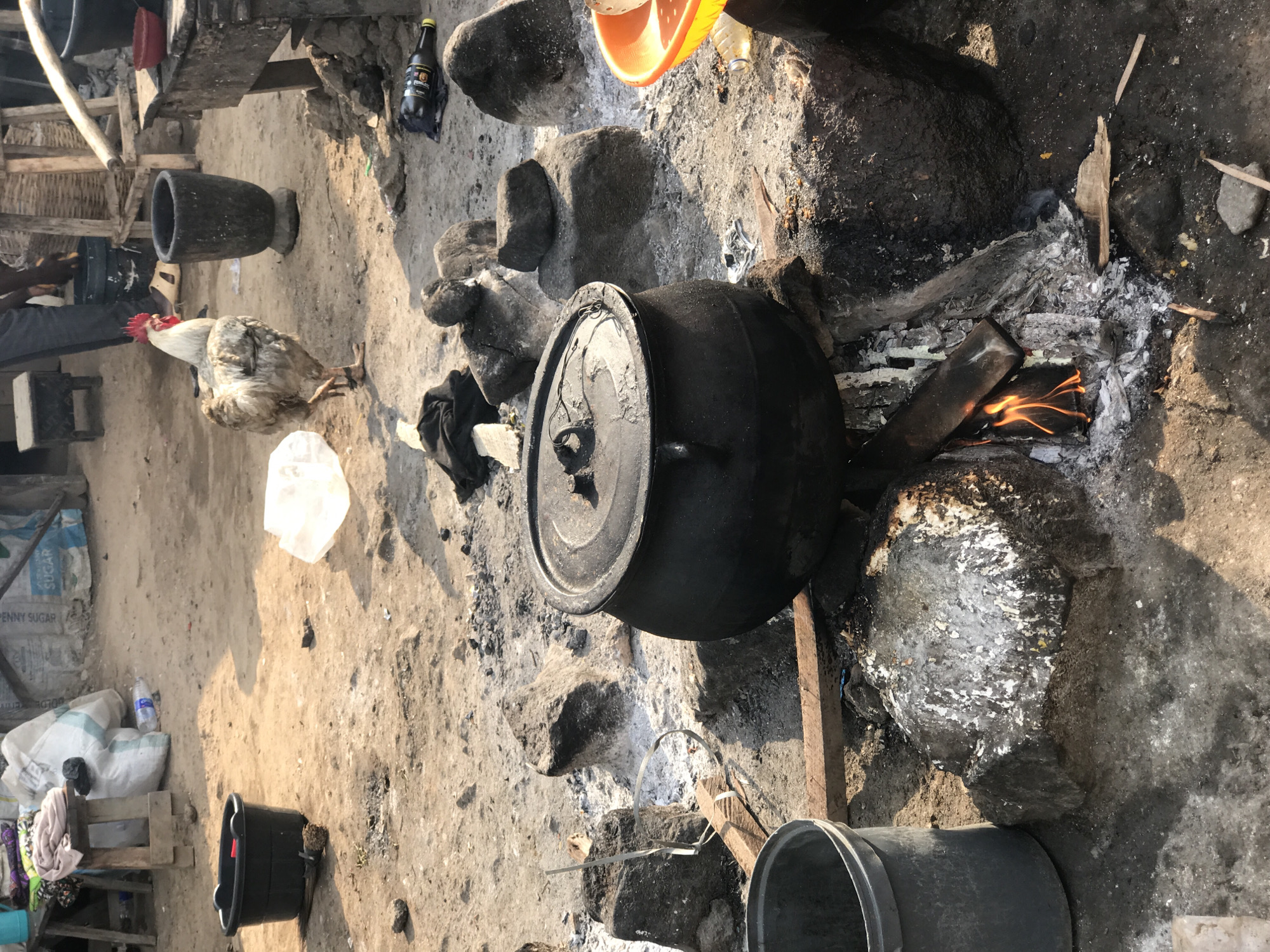 The image depicts a cooking scene in an outdoor setting in Dutsen Alhaji, Nigeria. A large, black pot with a lid sits atop a fire built with wood and stones. The fire is actively burning with visible flames. The surrounding area is covered in ash and soot. A chicken stands near a mortar, to the left of the fire, and a bucket is in the lower left corner. In the background there are bags and miscellaneous items.