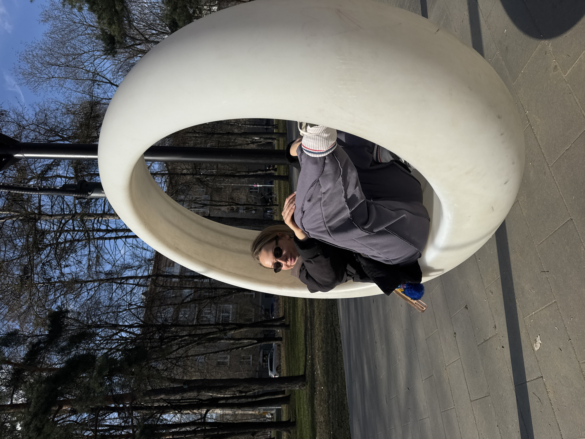 A woman is sitting inside a large, white, spherical swing in what appears to be a park or public space. She is wearing sunglasses, a black top, and a grey jacket, and a striped hat. The swing is suspended from a black metal frame. The ground is paved with grey bricks, and trees without leaves and buildings are visible in the background. The image appears to be taken during the daytime. Based on the geographical location determined from the image metadata, the location is Moscow, Russia.