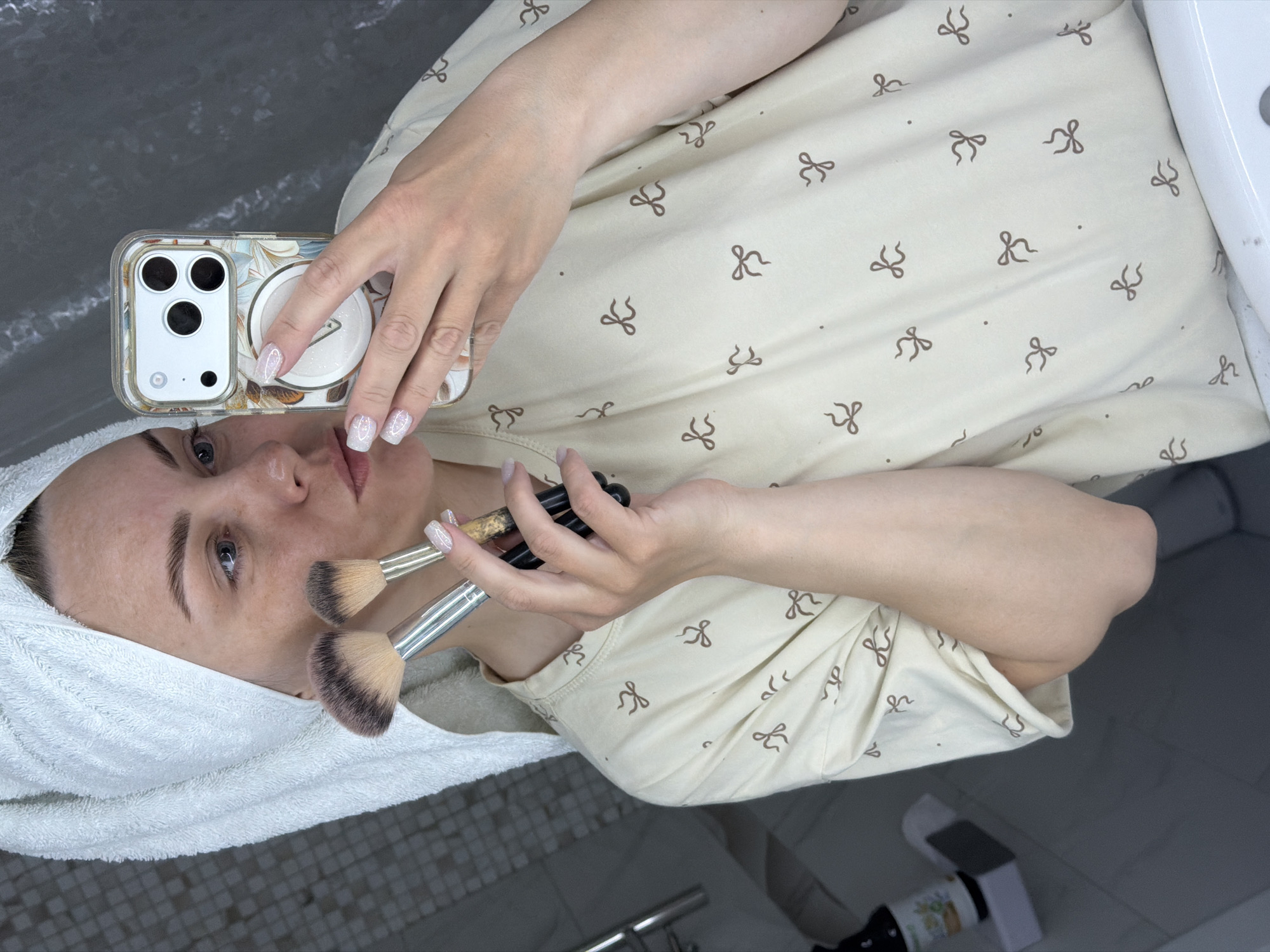 Here is a description of the image:
A woman is taking a selfie while holding makeup brushes. She is lying down and wearing a beige top with brown bows printed on it. A white towel is wrapped around her head. She is holding a white smartphone with a floral case. In her other hand, she holds several makeup brushes with silver and wooden handles and black bristles. The background appears to be a bathroom setting with grey tiles visible. The overall setting looks like a personal or home environment. No identifiable landmarks suggest that the image was taken in Votkinsk, Russia.