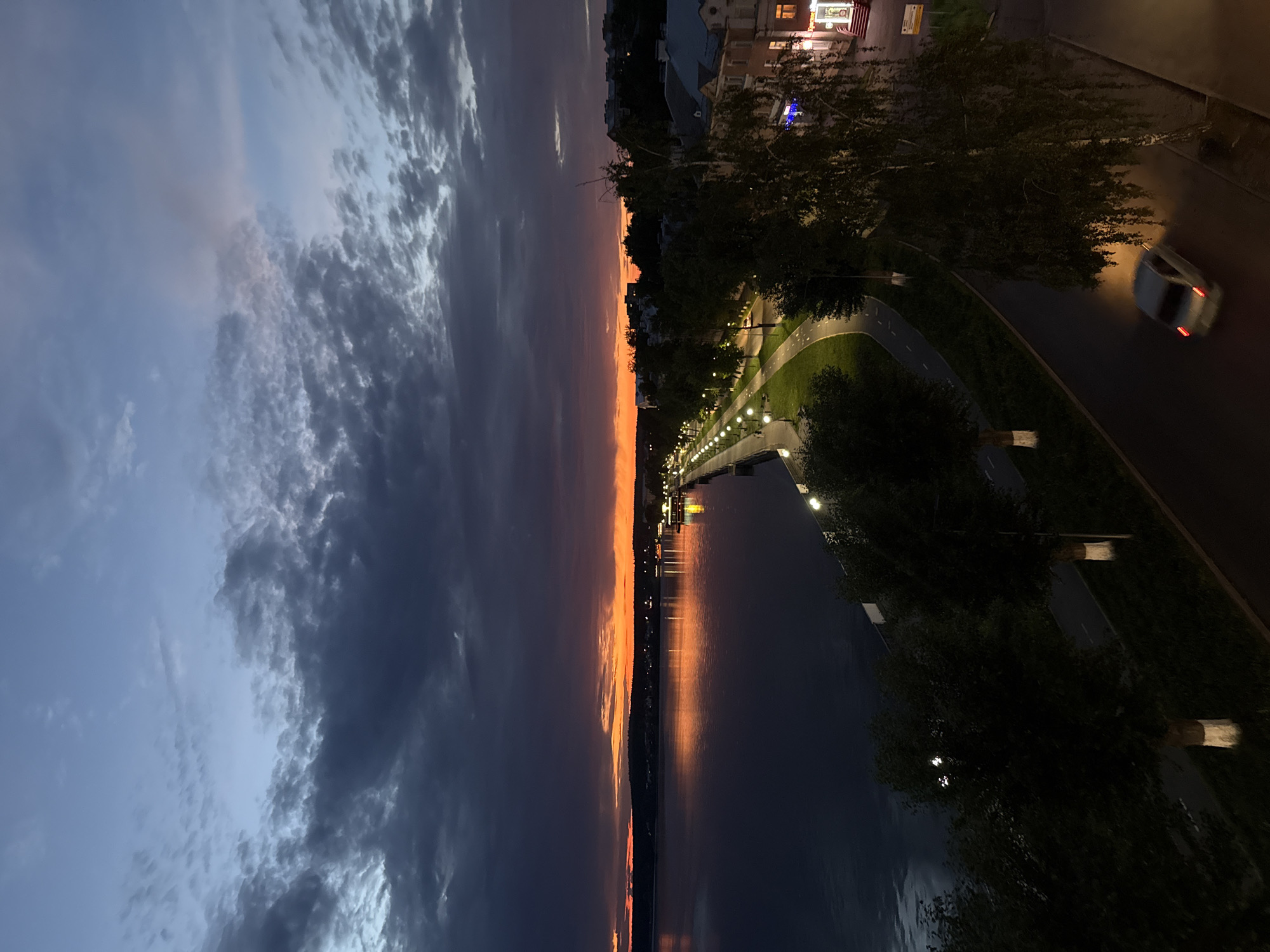The image captures a twilight scene along the Votkinsk Reservoir embankment in Votkinsk, Russia. The sky is a dramatic blend of dark, stormy clouds and the vibrant orange glow of a sunset near the horizon. The reservoir itself reflects the sky's colors, appearing as a dark, rippling expanse.
Along the embankment, a paved pathway is illuminated by a series of streetlights, creating a warm glow on the path and surrounding grass. Trees are planted along the pathway, some with their trunks painted white, a common practice in Russian cities. Beyond the pathway, the edge of the city is visible, with buildings showing illuminated windows and street-level lights.
A solitary car, its taillights illuminated, is driving on a road adjacent to the pathway, appearing slightly blurred due to motion. The overall atmosphere is serene, characteristic of dusk in a city situated by a large body of water.