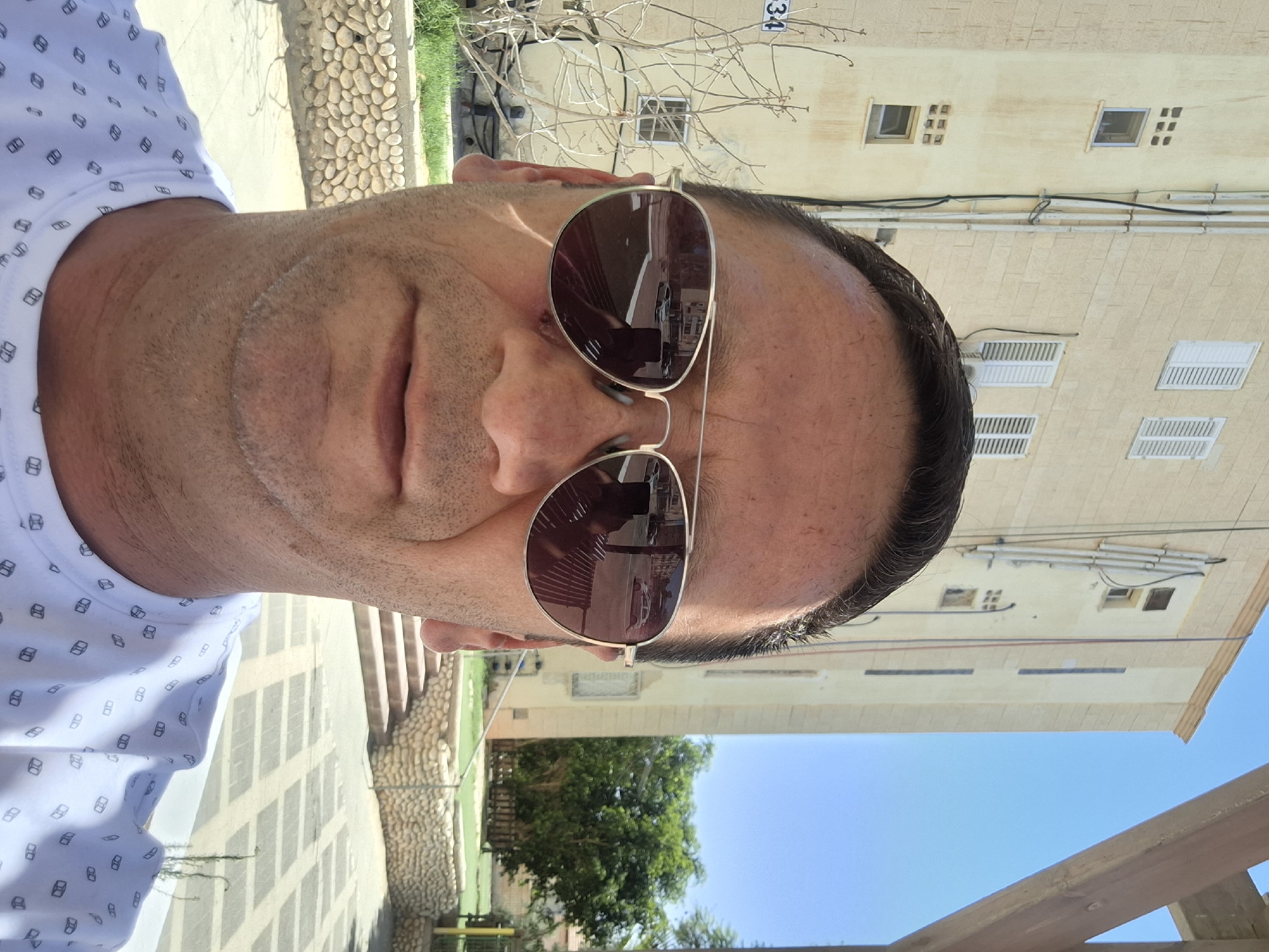 A man wearing aviator sunglasses and a white t-shirt with a repeating pattern of dark cubes is pictured in a selfie. He has short dark hair and stubble on his chin. The background shows a light-colored building with several windows and some external pipes. To the left of the building, a street sign reading "34" is visible on a post next to a bare tree. In the foreground, a low wall constructed of light-colored stones is present, with a paved area beyond it. The scene appears to be outdoors in a sunny climate, consistent with Yerocham, Israel. Reflections in the man's sunglasses show cars and a bench.