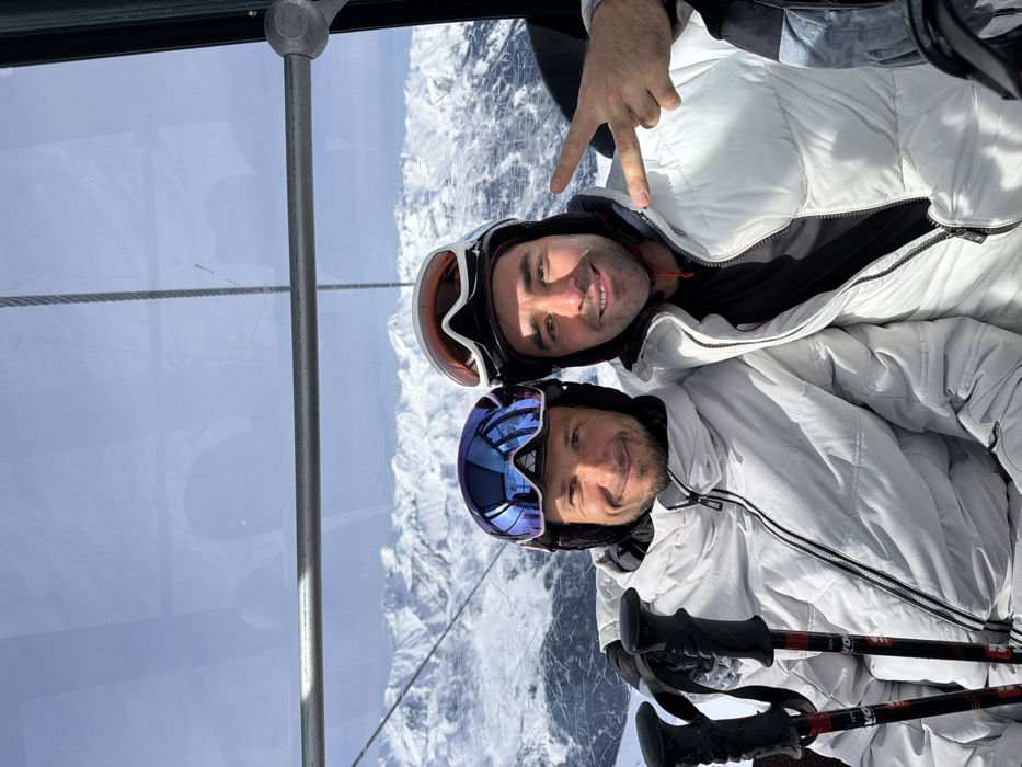Two adult males are depicted inside a ski gondola or cable car, presumably in the mountainous region of Les Allues, France, under clear daytime conditions. Both individuals are seated and looking towards the camera with smiles. The male on the left wears a dark ski helmet with blue reflective goggles pushed onto the helmet, and a light grey or white puffer jacket. Ski poles with black shafts and red accents are visible to his left. The male on the right wears a dark ski helmet with orange and white goggles pushed onto the helmet, and a white puffer jacket which is partially unzipped, revealing a black garment underneath. His right hand is raised, displaying a two-fingered "peace" gesture. The background, viewed through the clear windows of the cabin, consists of extensive snow-covered mountain ranges under a bright, clear sky. A horizontal metal bar and vertical support structure from the gondola interior are visible in the upper portion of the frame.