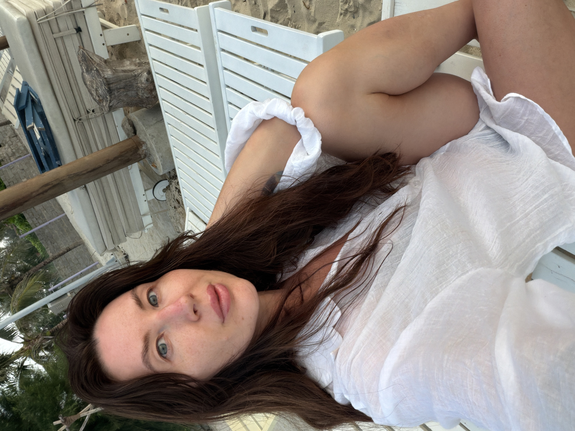 A woman with long, dark brown hair and blue-green eyes is reclining on a white lounge chair on a sandy beach. She is wearing a loose, white, sheer shirt. Her right arm is bent and resting on her side. In the background, there are more white lounge chairs, palm trees, and what appears to be a wooden pole supporting a canopy structure. The scene is outdoors, suggesting a tropical or beach resort setting. The presence of lounge chairs indicates a place of relaxation and leisure. The lighting suggests it might be late afternoon or early morning. The specific location in Dong Ninh Hoa Ward, Vietnam, is not visually evident from the image itself, but it suggests a coastal area within that region.