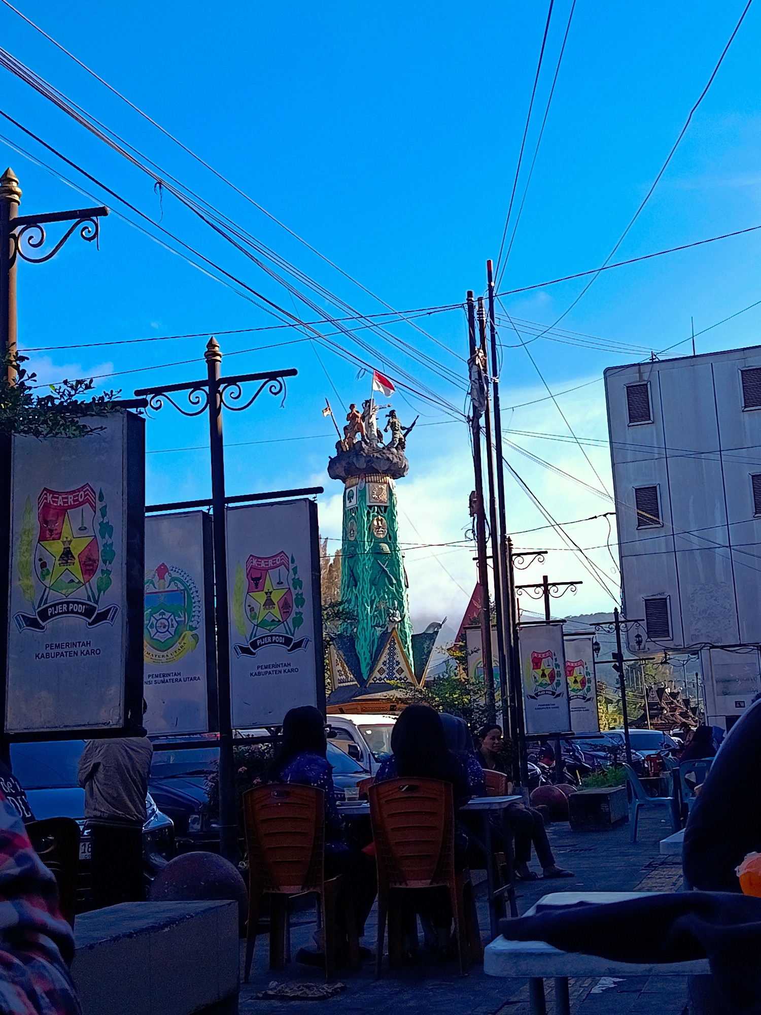 The image captures a street scene in Berastagi, Indonesia, under a bright blue sky with a clear view of overhead power lines. In the foreground, several people are seated at tables outside, with some appearing to be dining. The focus of the scene is a tall, ornate monument in the background, topped with figurines and a flag. Several banners are displayed along the street, featuring emblems and text that read "PEMERINTAH KABUPATEN KARO" and "PIJER PODI," indicating a connection to local government. The environment appears to be an urban setting, with buildings visible and vehicles parked along the roadside. The lighting suggests it is daytime, with the sun casting shadows and illuminating the scene.