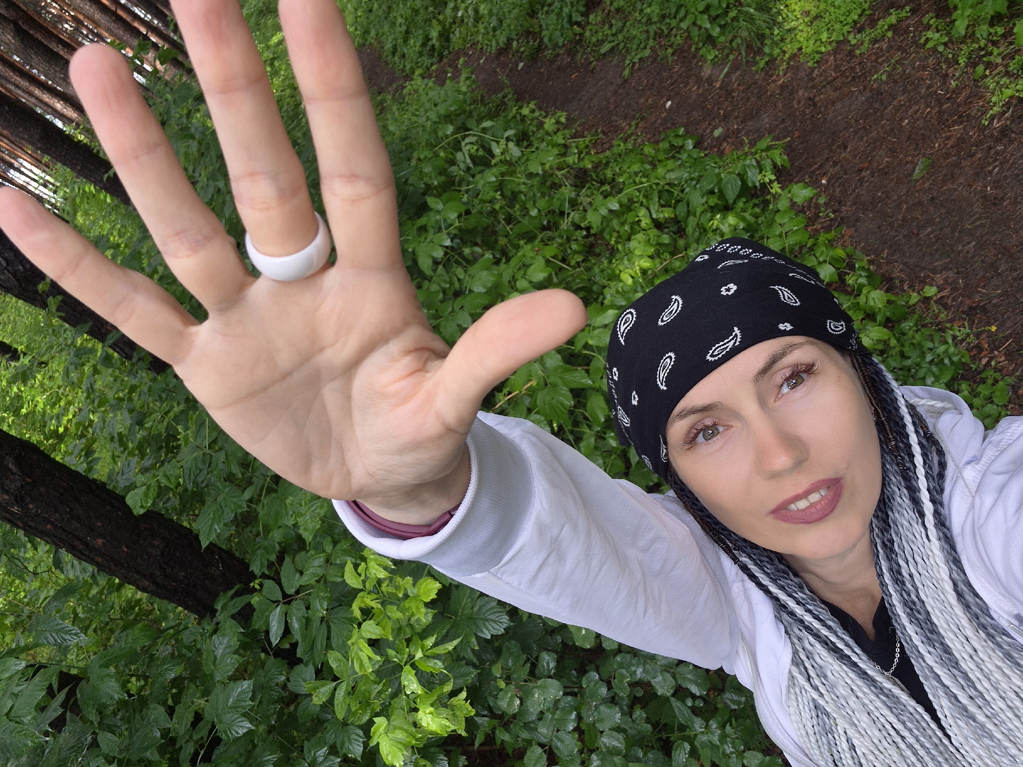 A woman is taking a selfie outdoors in a forest. She is wearing a black bandana with a paisley pattern, a white jacket over a black shirt, and has braided hair in shades of gray and white. She is wearing a white ring on her left index finger. Her right arm is extended towards the camera, with her palm facing forward. The background is a dense forest with green foliage and trees. A dirt path is visible to the right of the woman. The image appears to be taken during the daytime, with natural lighting. The location is identified as the city of Chaikovsky, Russia.