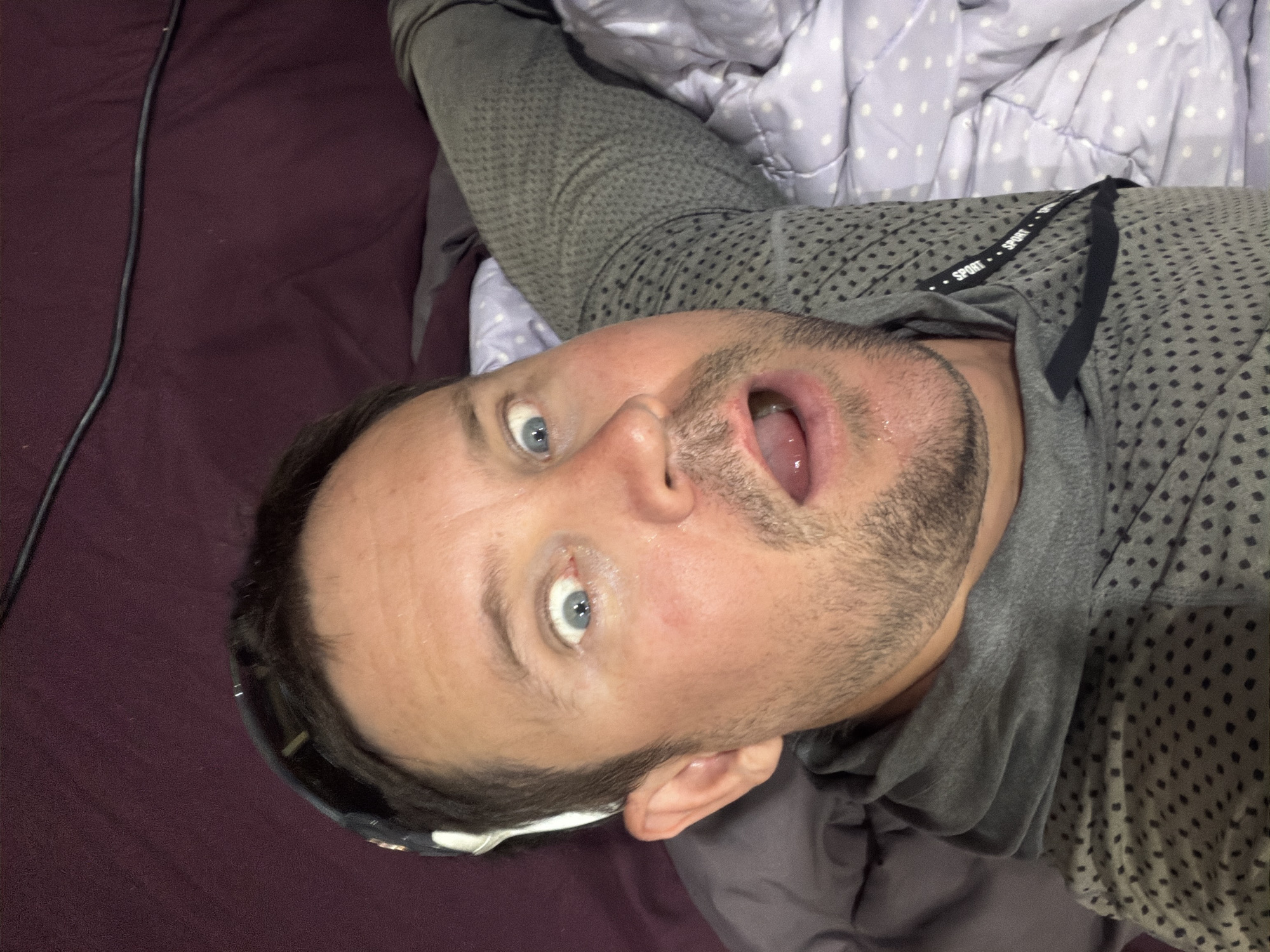 A man is lying down, looking up at the camera with a surprised expression. He has blue eyes, brown hair, and a beard. He is wearing a gray hoodie and has sunglasses on his head. The background is a dark purple fabric, possibly a blanket or bedspread. There is also a white and purple polka-dotted blanket visible. The scene appears to be indoors, possibly in a bedroom. There is no discernible information about the location in Baan Mae Nam, Thailand.