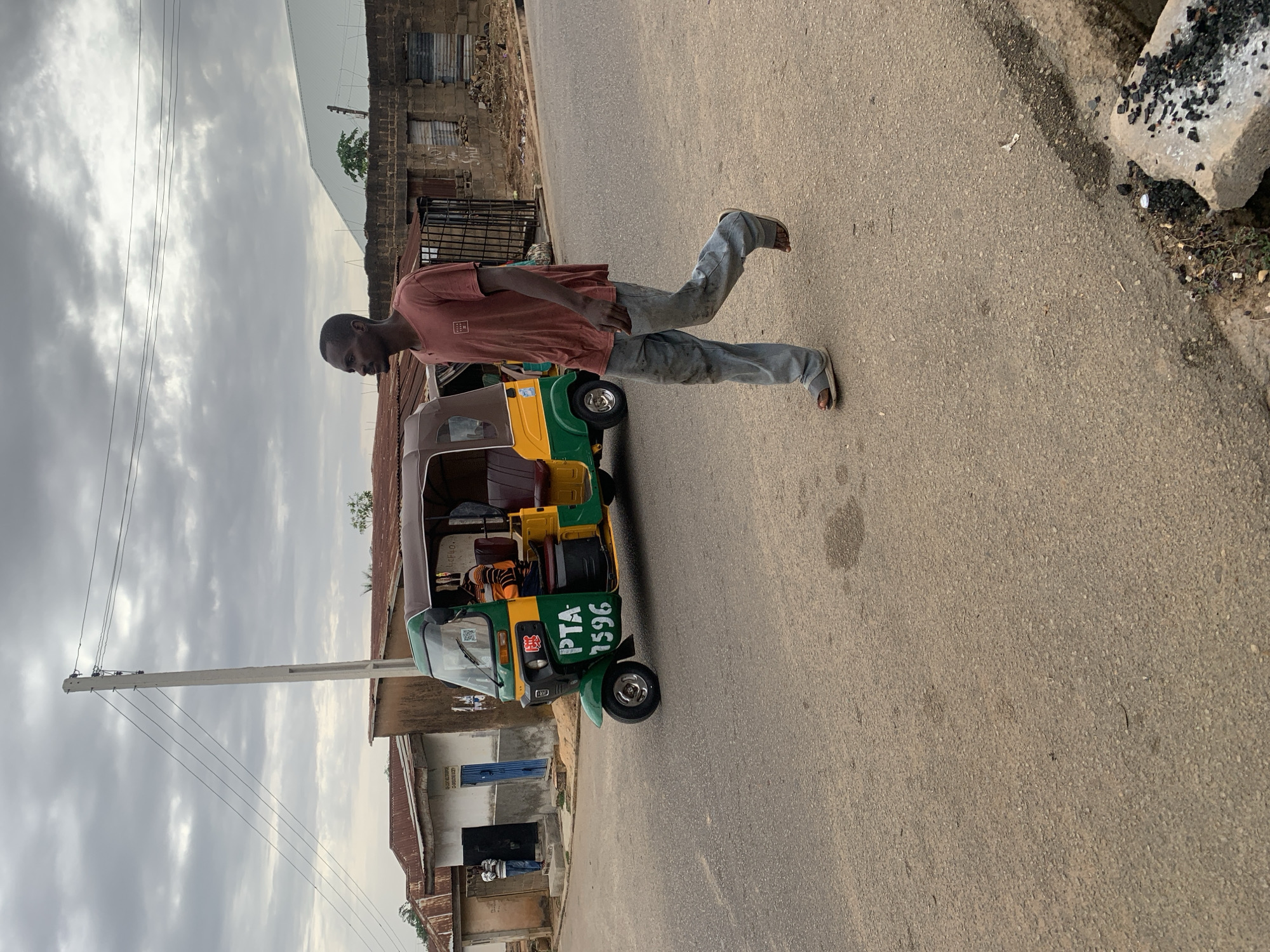 The image captures an everyday street scene, likely in Jos, Nigeria, featuring a man, a three-wheeled vehicle, and urban infrastructure under an overcast sky.
In the foreground, a man is walking on an asphalt road. He is wearing a reddish-brown short-sleeved shirt, light blue jeans, and sandals. His body is in mid-stride, with his left leg bent and raised, and his head slightly lowered, looking forward or down.
Adjacent to the man, on his right, is a green and yellow three-wheeled auto-rickshaw, commonly known as a "keke." The vehicle has a brown canopy and a visible front wheel. Prominently displayed on its side in white lettering is "PTA 7596", likely an identification number or license plate. A red emblem is also visible near the front of the keke.
The street itself is paved with asphalt and shows some wear. Along the right edge of the road, there's a concrete structure, possibly a curb, covered with dark debris that resembles charcoal or ash.
The background consists of low-rise buildings constructed with concrete blocks and topped with corrugated iron roofs, indicative of typical residential or commercial structures in the region. To the left, a tall, light-colored utility pole with several power lines stretches into the cloudy sky. The sky is largely covered with dense, grey clouds, suggesting an overcast day. A small, green tree is visible in the distance among the buildings on the right. The overall impression is one of a functional urban setting with local transportation.