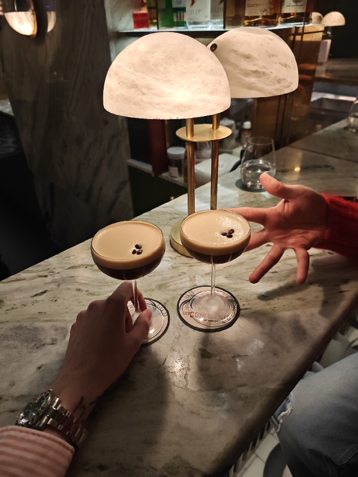 The image captures a close-up, eye-level view of two espresso martinis on a polished marble bar counter, illuminated by ambient lighting. The setting appears to be an upscale bar or lounge during the evening.
In the foreground, two espresso martinis are presented in classic stemmed martini glasses. Each glass contains a dark brown liquid topped with a creamy, light brown foam, and garnished with three coffee beans arranged in a triangular pattern on the foam. The glass on the left is being held by a hand with a prominent gold-toned metal watch with a fluted bezel on the wrist. The person's sleeve, visible on the left, is striped in white and a light color, possibly pink or peach. Beneath each martini glass is a round coaster. The coaster under the right glass clearly displays text that reads "LES CONTINENTS" and "DE COMORES," along with a stylized circular design.
To the right of the martinis, another hand, with fingers spread, is positioned near the right cocktail. This hand appears to belong to someone wearing a vivid red long-sleeved garment, possibly a sweater. A glimpse of blue denim trousers is visible at the bottom right corner of the frame.
Dominating the mid-ground and providing a warm glow are two decorative table lamps with unique, dome-shaped shades made of a translucent, milky white material that has a swirled or veined texture, resembling alabaster or marble. These shades are supported by slender brass-colored stands.
The bar counter itself is made of a light-colored marble with distinct green and grey veining. In the blurred background, several bottles of liquor are visible on shelves, and a clear glass of water sits on the counter behind the lamps, further suggesting a bar environment. The overall lighting is dim and warm, creating an intimate and sophisticated atmosphere.