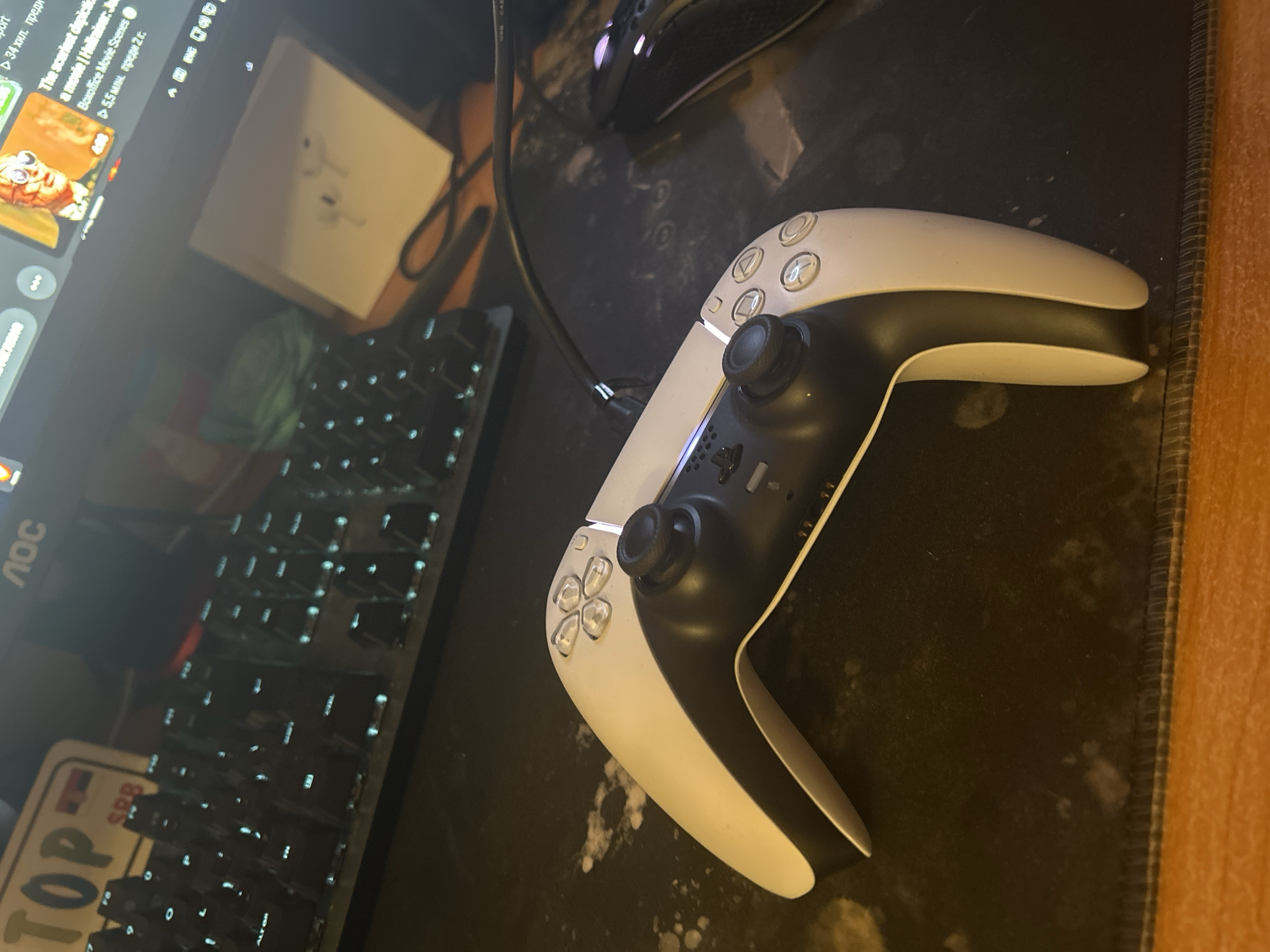 The image presents a desk setup. Centered is a white PlayStation 5 DualSense controller with a USB cable attached. Adjacent to the controller is a black computer mouse. To the left, a black mechanical keyboard is visible, partially covered by a license plate souvenir with "TOP" written on it, and the letters "SPB". Behind the keyboard, an opened box of what appears to be Apple AirPods can be observed. The monitor, an AOC model, displays a YouTube video, recognizable by the play button, title, and timestamp. The desk is covered with a black mousepad, bordered with a brown wooden edge on the right side. There are no people in the image. The photo lacks information that would place the location within Sofia, Bulgaria.