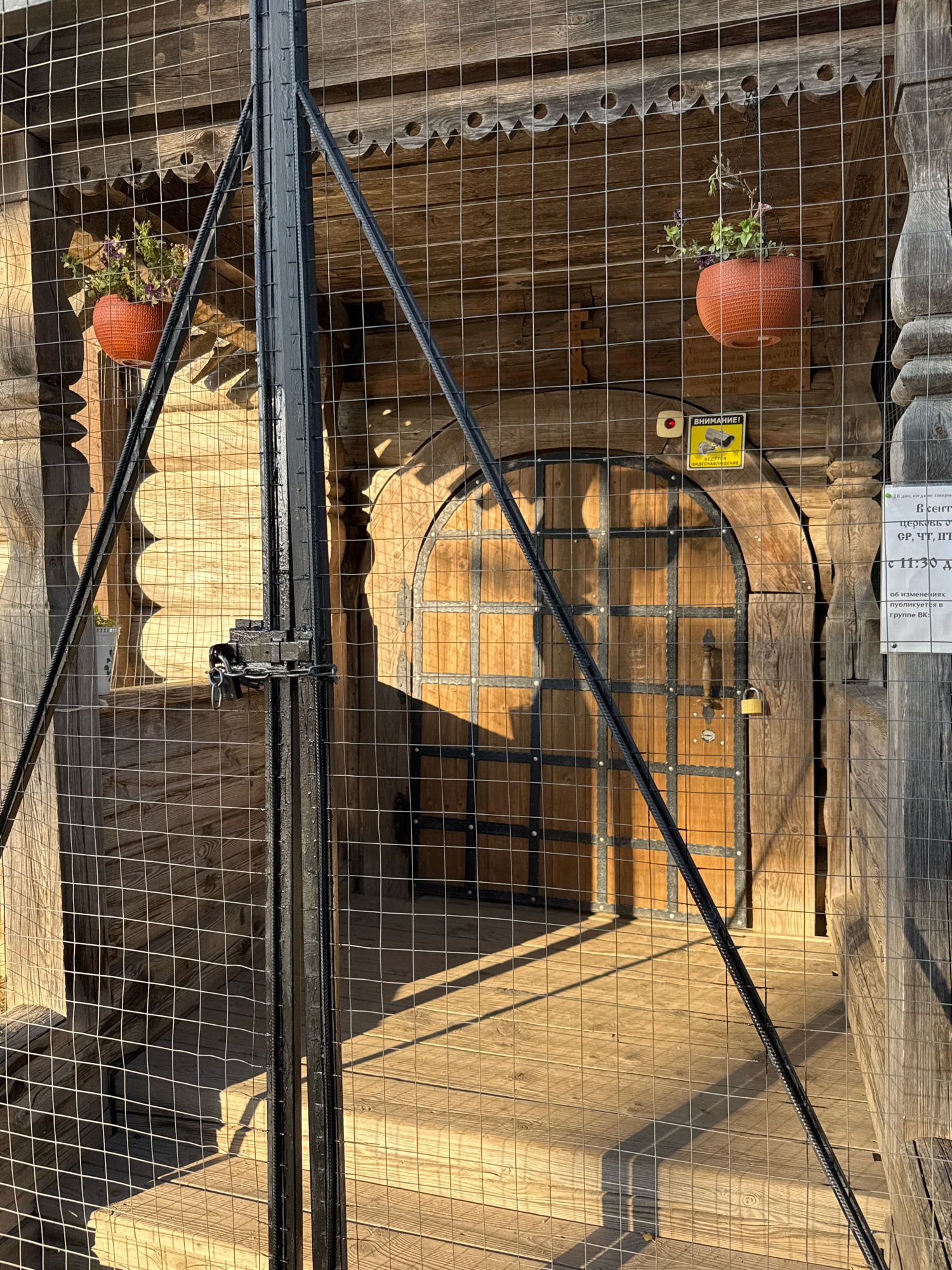 This image shows a locked gate in front of a wooden door at a wooden structure in Torzhok, Russia. The gate is made of metal bars and wire mesh and is secured with a large padlock. The wooden door is arched and has metal accents. There are two hanging flower pots with plants, one on each side of the door. A yellow warning sign with a camera icon and the word "ВНИМАНИЕ!" (Attention!) is attached to the wall above the door. The scene appears to be outdoors, with sunlight casting shadows on the wooden planks of the walkway.