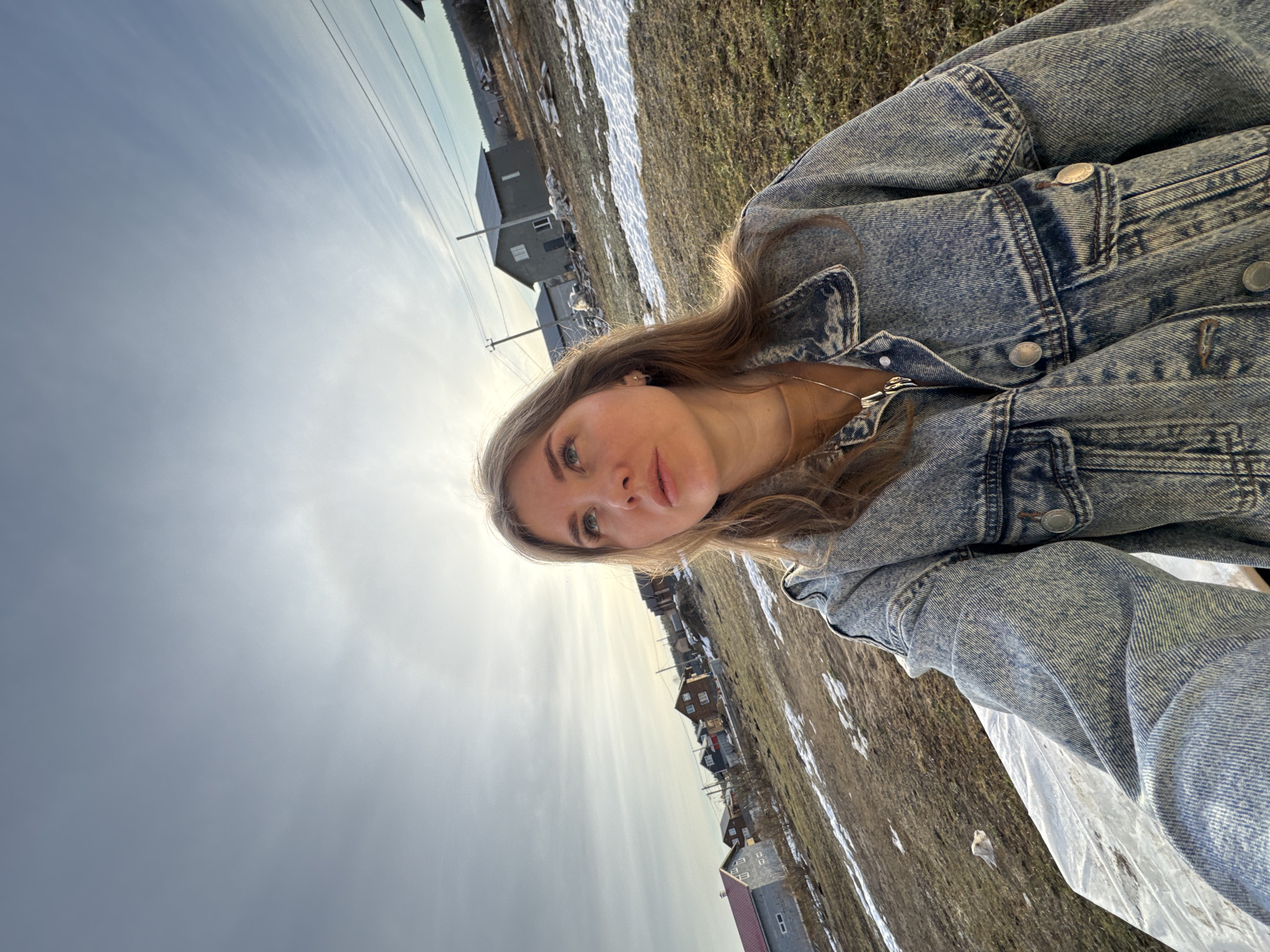 A woman is taking a selfie outdoors in Gornyi (Dvurechenskoe S/P), Russia. She is wearing a denim jacket and a necklace. The background shows a landscape with patches of melting snow, grass, and houses. Overhead power lines are visible in the sky. The sky is overcast.