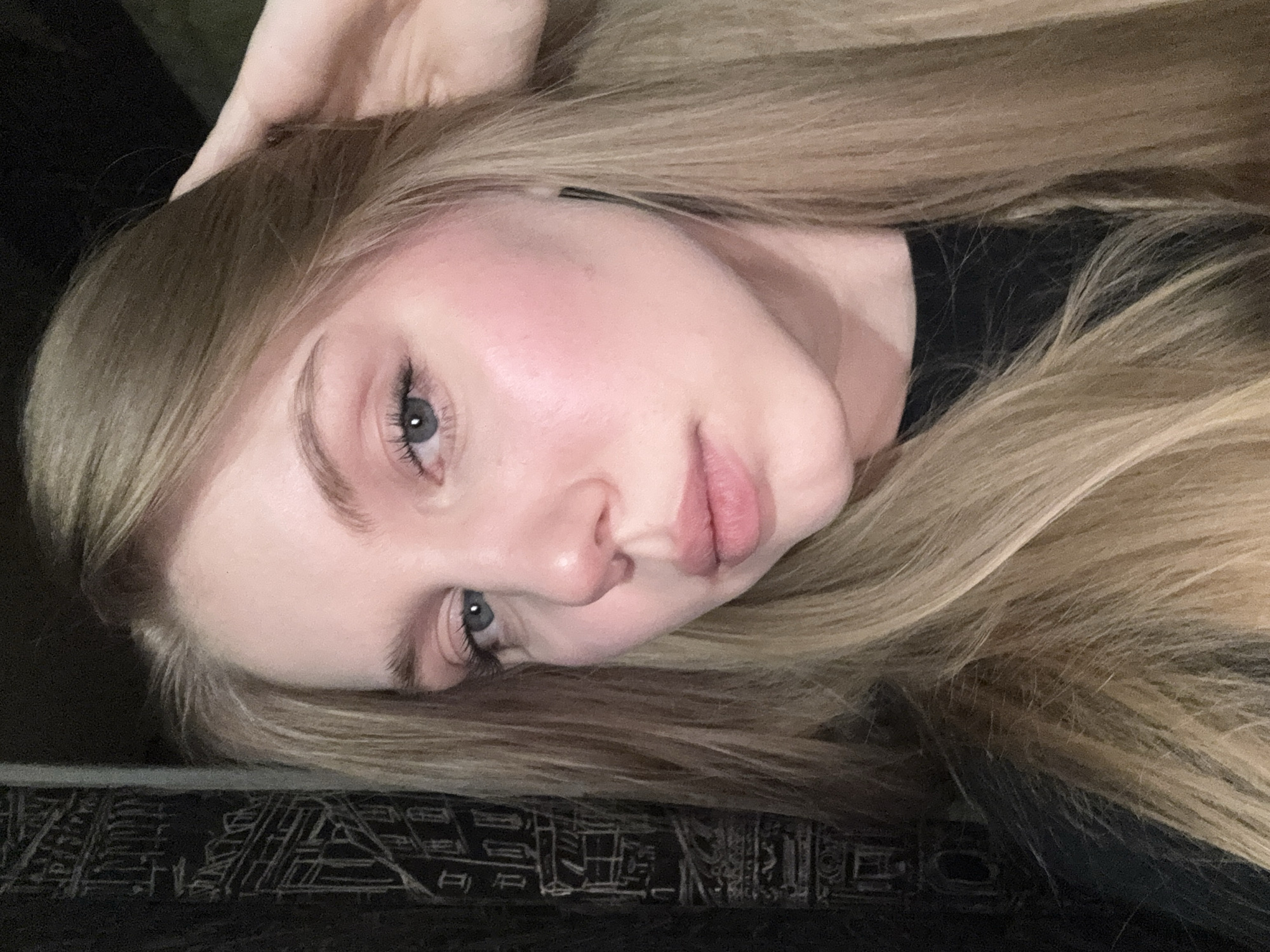 The scene depicts a close-up portrait of a young Caucasian female in Perm, Russia. She possesses long, straight blonde hair that frames her face and extends beyond her shoulders. Her skin is fair, with a noticeable pink flush on her cheeks. She has blue eyes and light-colored eyebrows. Her lips are light pink and slightly parted. She is looking directly at the viewer, with her head subtly tilted. Her right hand (viewer's left) is partially visible, with fingers appearing to rest on or manipulate her hair near her temple or ear. She is wearing a dark, likely black, top. The background is predominantly dark and out of focus. To the left of the subject, a pattern resembling architectural outlines of buildings, rendered in a lighter tone against the dark background, is visible. The lighting is soft and appears to originate from the front, illuminating the subject's face.