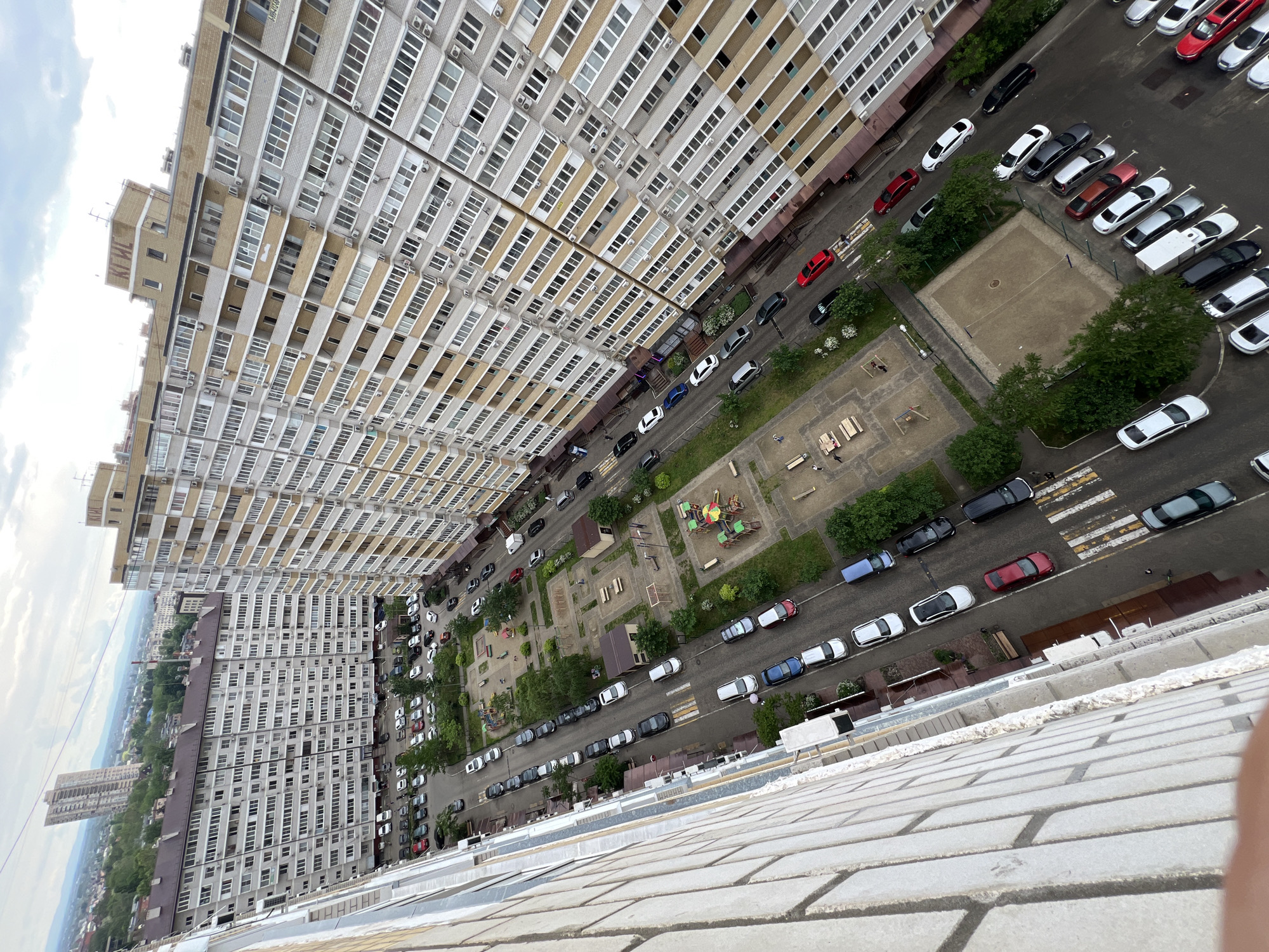 The image shows an aerial view of a residential area, likely taken from a high-rise building. There are multiple large apartment blocks with rows of windows and air conditioning units. Streets run between the buildings, filled with parked cars of various colors. There are playgrounds and a volleyball court visible amidst the buildings.
Based on the architectural style and the overall layout, this scene is potentially located in the city of Krasnodar, Russia.