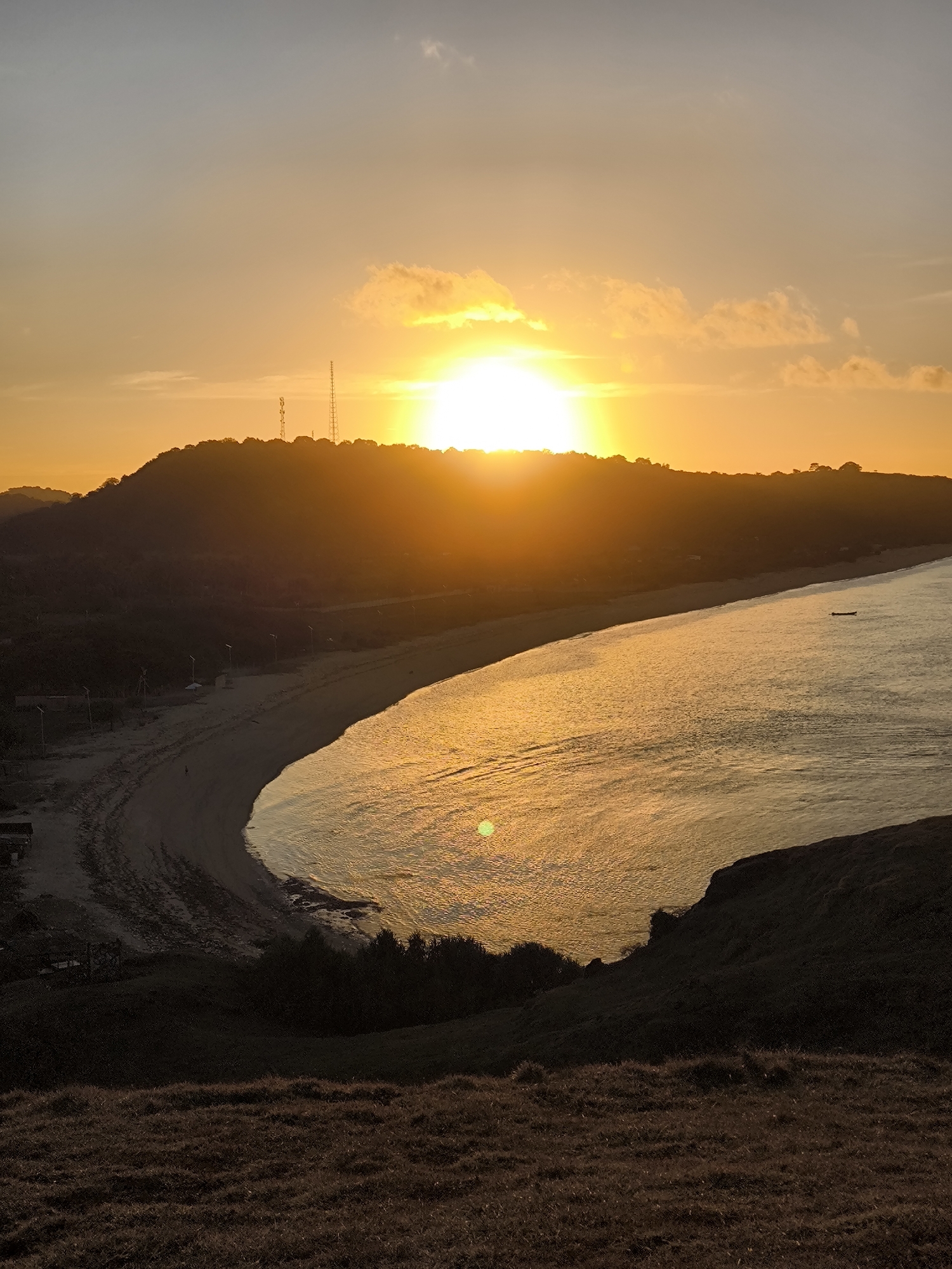 The image shows a sunset scene in Kuta, Indonesia. The sun is setting behind a hill, casting a golden glow over the bay. A long, curving beach is visible along the shoreline. The water is calm and reflects the sunlight. On the hill there are tall structures that resemble towers. The foreground consists of grassy hillsides. A small boat can be seen on the water.