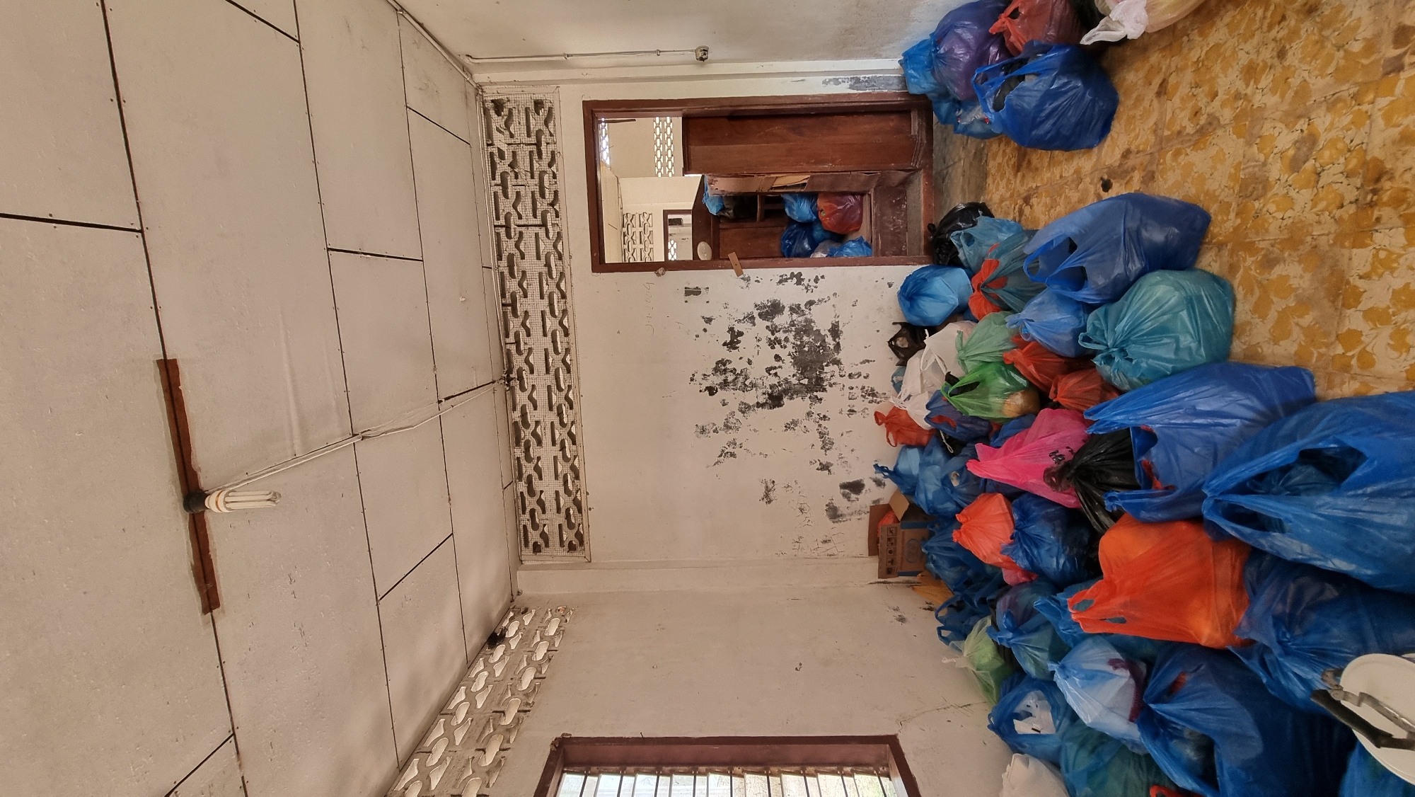The image depicts a cluttered room, likely a storage area, filled with numerous plastic bags of various colors, predominantly blue, orange, pink, and green. These bags are piled high on the floor, creating an obstacle course. A wooden doorway on the right side of the image leads to another room, also containing a substantial number of similar bags. The walls are dirty and peeling in places, and the ceiling appears to be made of corrugated material with exposed wiring and a light fixture. A window with slatted blinds is visible on the left. The floor is tiled with a yellow and brown pattern. The scene suggests a donation or distribution point, possibly related to disaster relief or charity work, within the city of Tumpokbeurandang, Indonesia. There are no people visible in the image.