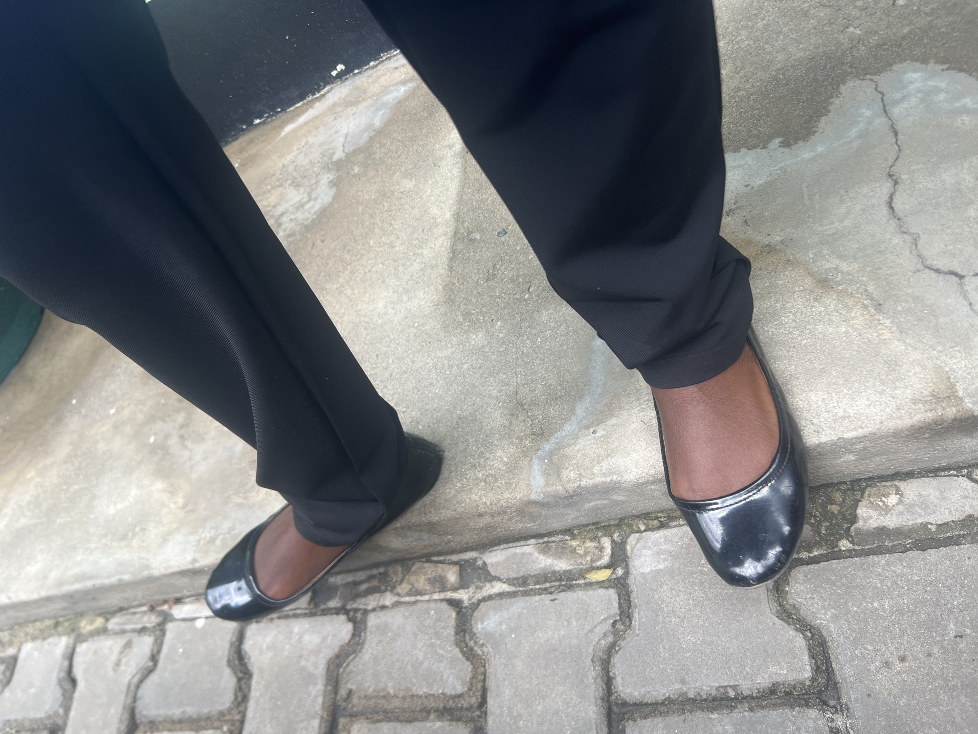 The image captures a ground-level view of a person standing outdoors, focusing on their lower legs and feet. The subject is wearing black trousers, which appear to be made of a smooth fabric, and black patent leather ballet flats. The person's skin tone, visible around the ankles and feet within the shoes, is dark brown.
The setting is an outdoor paved area, likely a sidewalk or urban pathway. The ground consists of two main sections: a smoother, light grey concrete surface and a section of grey interlocking pavers. Some cracks and minor stains are visible on the concrete. The lighting suggests it is daytime, with bright conditions, and subtle shadows indicating sunlight. No specific weather conditions can be inferred beyond it being clear. There is no visible text in the image. Given the context, the location is Dar es-Salaam, Tanzania.