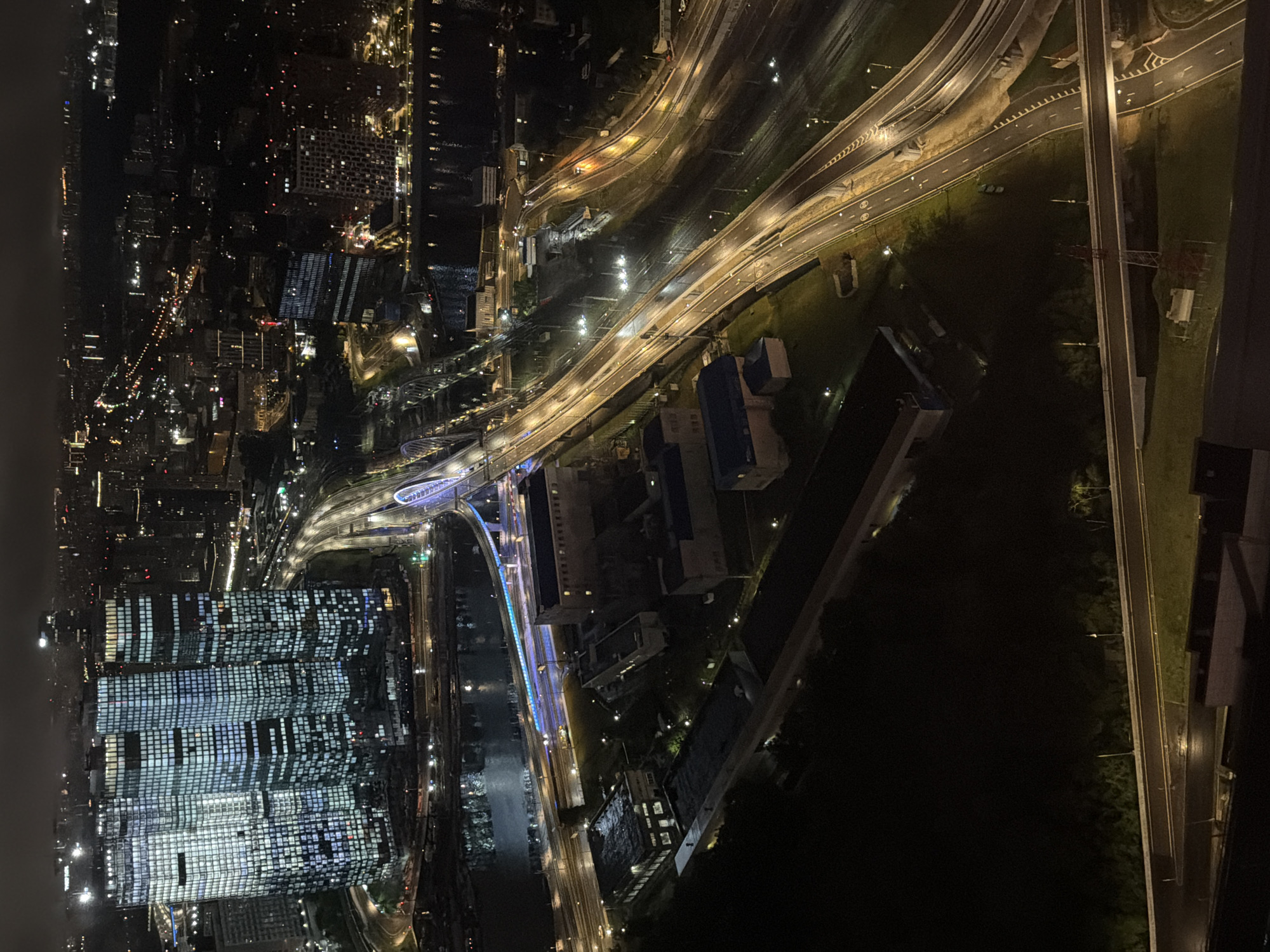 This aerial nighttime image captures a sprawling urban landscape in Khimki, Russia, dominated by illuminated skyscrapers and a complex network of roadways. In the foreground, a body of water reflects the city lights, bordered by industrial buildings and a multi-level highway system. The highway, with its bright streetlights, curves and intersects, showcasing the city's infrastructure. Beyond this, several modern skyscrapers rise impressively, their facades covered in a grid of glowing windows, suggesting a vibrant business district. The background fades into a less defined cityscape, with more distant lights indicating a vast urban sprawl under a dark, cloudy sky. The scene is devoid of visible human activity, presenting a static yet dynamic portrayal of urban life at night.