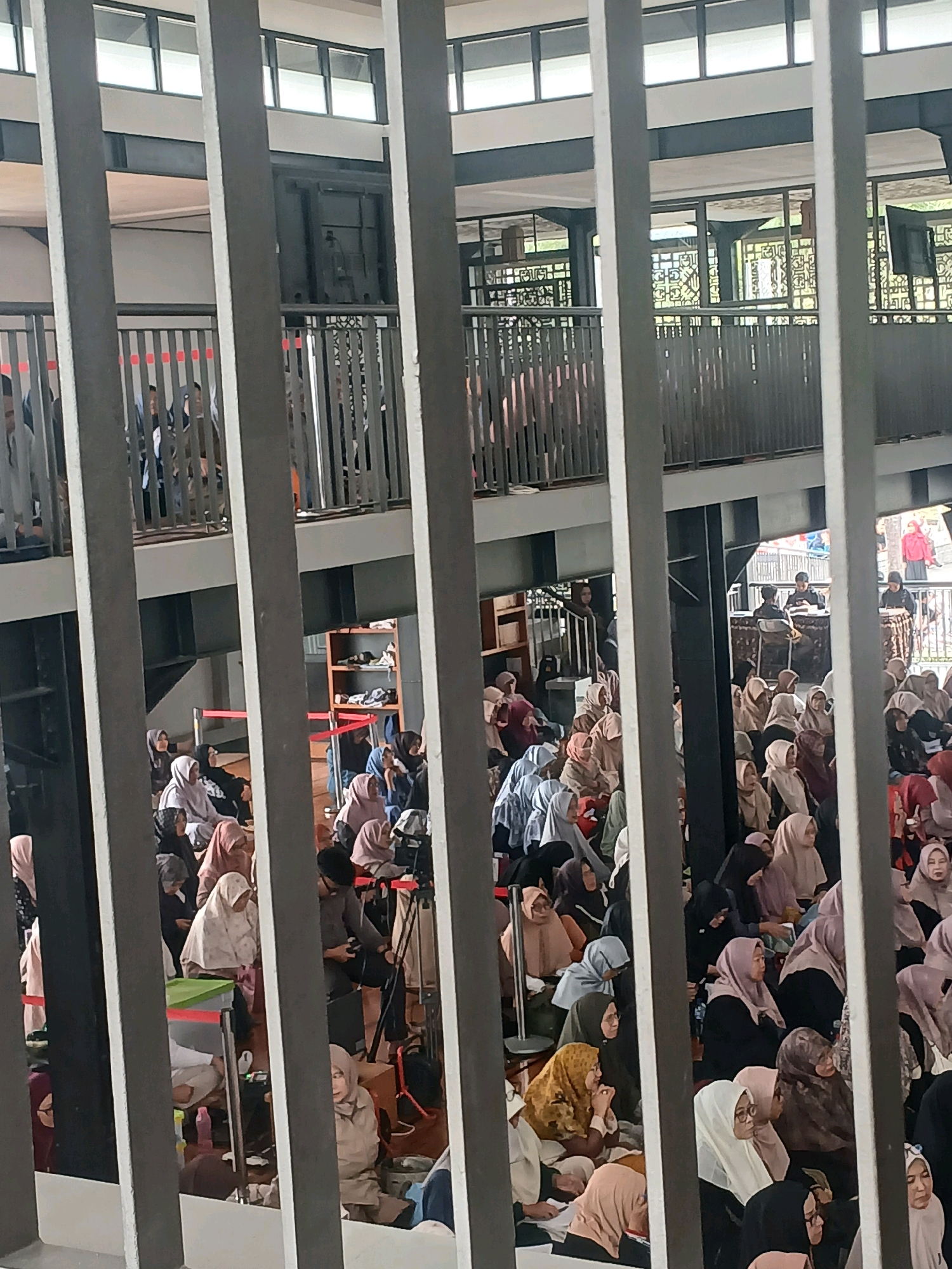 The image captures an indoor gathering of a large group of people, predominantly women wearing hijabs, seated on the floor. The scene appears to be a public space with architectural elements suggesting a modern building, possibly a community center or place of worship. The perspective is from behind a set of thick, vertical metal bars, creating a layered view of the space.
In the foreground, the metal bars dominate, partially obscuring the view. Behind them, a crowd is visible, mostly seated in rows. Many individuals are wearing hijabs in various colors, including beige, brown, white, and black. Some are looking towards a central area, while others are interacting or looking down.
Further in the background, there are elevated walkways and balconies with railings, where more people can be seen, some standing and others sitting. The architectural style includes exposed structural beams and large windows. Shelving units are visible in the mid-ground, displaying shoes and other items. A few individuals in the background appear to be engaged in an activity at a table, possibly a presentation or discussion. The lighting suggests an indoor environment, possibly illuminated by artificial lights. The overall impression is of an organized event or meeting. The city of Arjasari, Indonesia, is mentioned as the location, implying a cultural context where such gatherings are common.