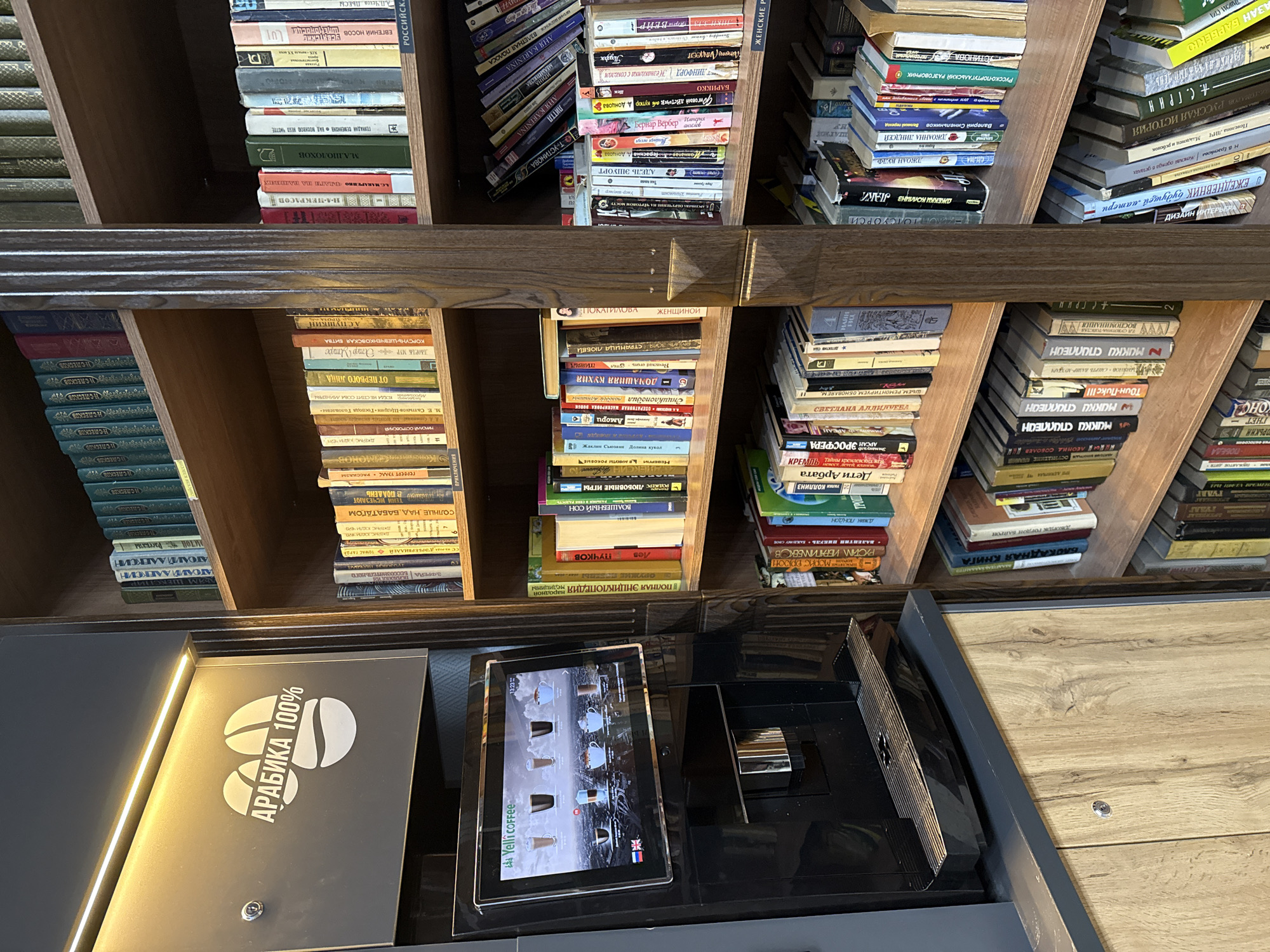 The image shows a section of a building interior, featuring a dark wood bookshelf densely packed with books. The books vary in height, color, and thickness, suggesting a diverse collection. The bookshelf is neatly organized. In front of the bookshelf is a dark counter with several devices on it. A tablet displays an image of coffee beverages, and a coffee machine is positioned next to it. The counter is illuminated with a strip of warm light. An inscription in Russian "АРАБИКА 100%" is visible on one of the components. Based on the text in Russian on the book covers and the presence of coffee-related items, the image is likely taken inside a coffee shop or library in Izhevsk, Russia.