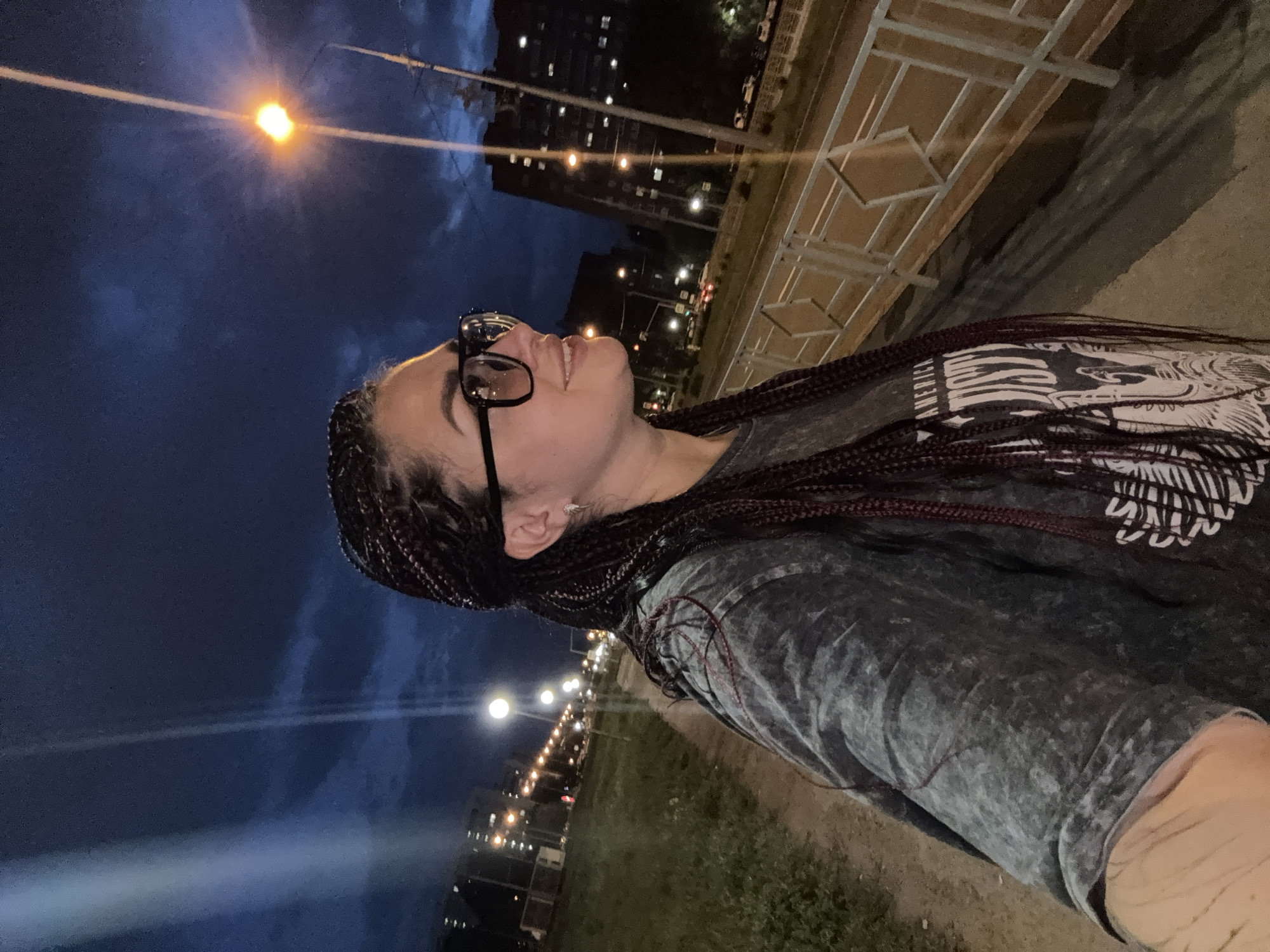 A young woman with dark braided hair and glasses is captured in a selfie at night, standing on a sidewalk next to a street. She is wearing a dark, marbled-patterned t-shirt with white graphic text. The background shows a dimly lit urban environment with streetlights, buildings, and a road with vehicles. The sky is dark blue with clouds, suggesting it's evening or night. The location is identified as Tolyatti, Russia, specifically along a pathway with a metal guardrail.