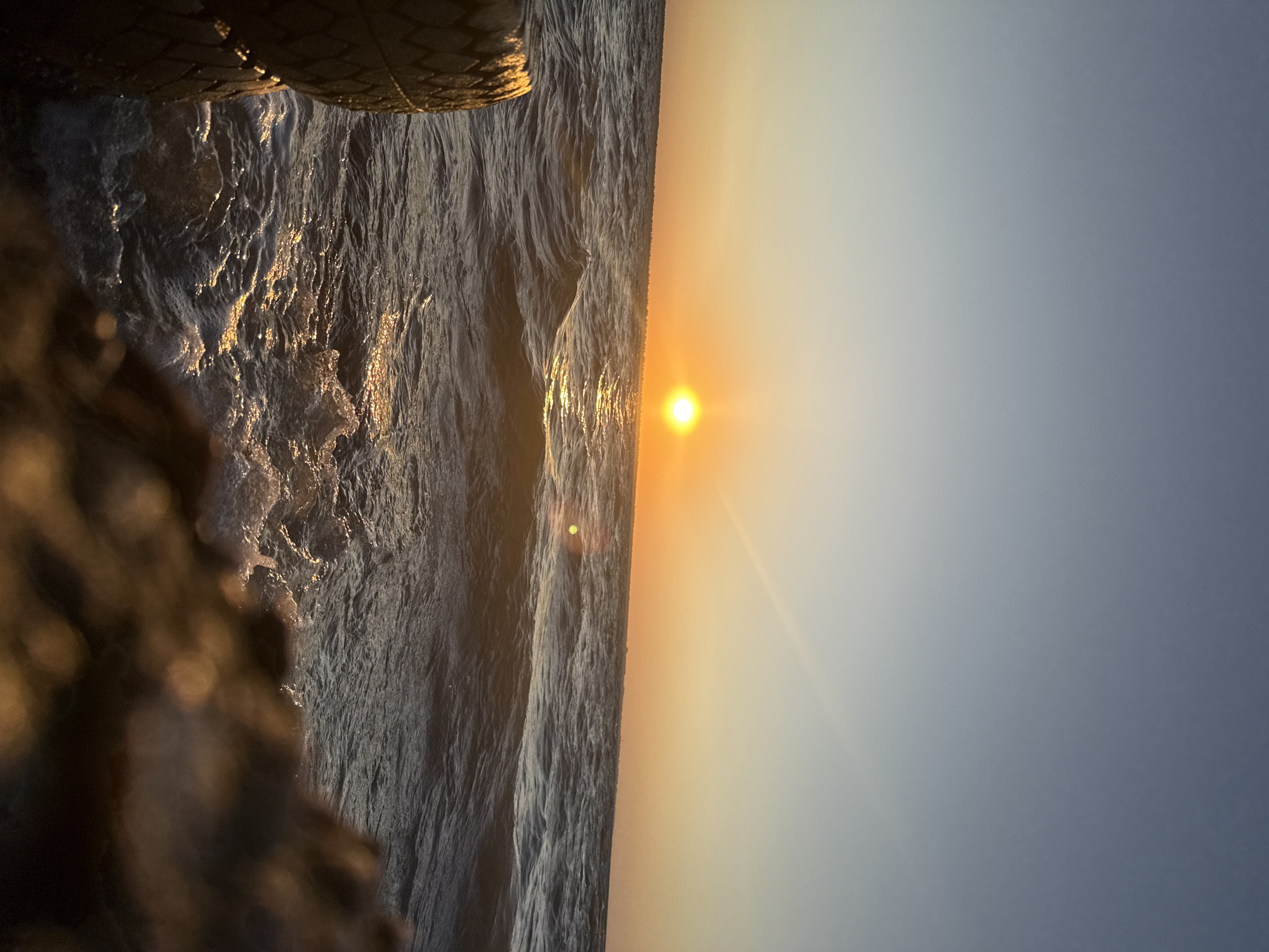 The image shows a serene sunset over the Black Sea, likely near Zatoka, Ukraine. The sun, a vibrant orange orb, is low on the horizon, casting a shimmering golden path across the choppy water. Gentle waves roll towards the shore, creating white foam as they break. In the foreground, the weathered texture of a tire is visible on the left, suggesting it may be part of a breakwater or a mooring. On the right, a blurred, rocky foreground adds depth to the scene. The sky transitions from a pale blue at the top to a warm orange glow near the horizon, indicating the approaching evening. No people are visible in the frame.