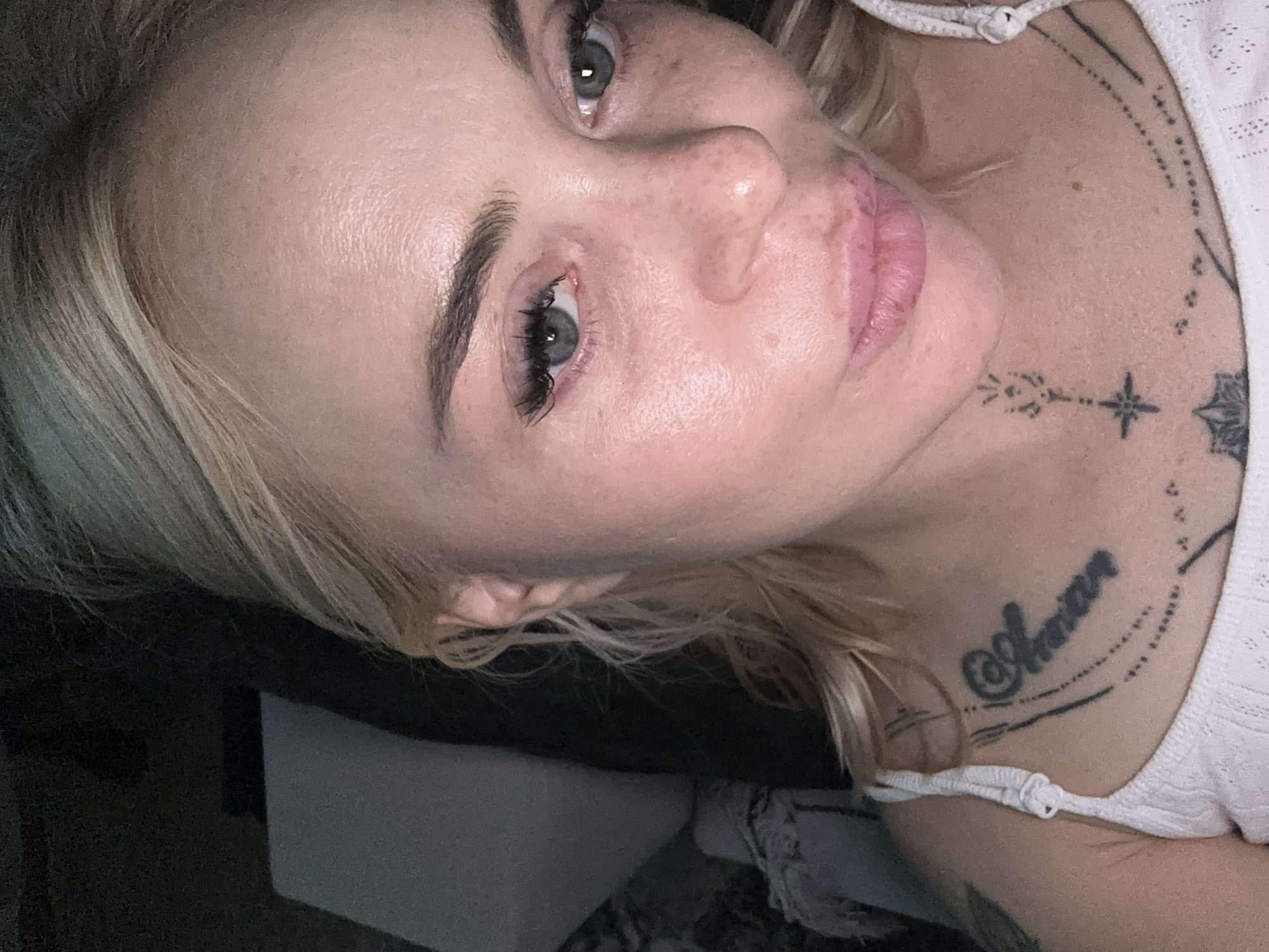 A close-up, overhead photograph depicts a fair-skinned individual with blonde hair and blue-grey eyes, appearing to be a woman in her 20s or 30s. Her face is directed towards the camera, illuminated by a frontal light source that highlights her forehead, nose, and cheeks. Visible skin texture includes pores, freckles across the nose and cheeks, and some areas of redness. Her eyebrows are dark and well-defined. Long, dark eyelashes frame her eyes, which have a slight reddish tint in the inner corners. Her lips are full, pink, and slightly parted. Her blonde hair is wavy and appears casually styled, with strands falling around her face and shoulders.
The individual is wearing a white, possibly knit or crocheted, spaghetti-strap top. Multiple tattoos are visible on her upper chest and neck. A prominent tattoo on her upper sternum features stylized script, possibly reading "Aman" or "Amara," accompanied by decorative lines and dot patterns. On her central throat, a linear tattoo resembling a small star or cross with vertical embellishments is present. Additional faint linear patterns are visible on her collarbone area.
The background is predominantly dark and out of focus, suggesting an indoor setting with low ambient light. A light grey object, possibly a pillow or furniture, is partially visible on the left side of the frame. The image captures a direct gaze and a neutral to slightly pouting expression.
Location: Unknown, Unknown.