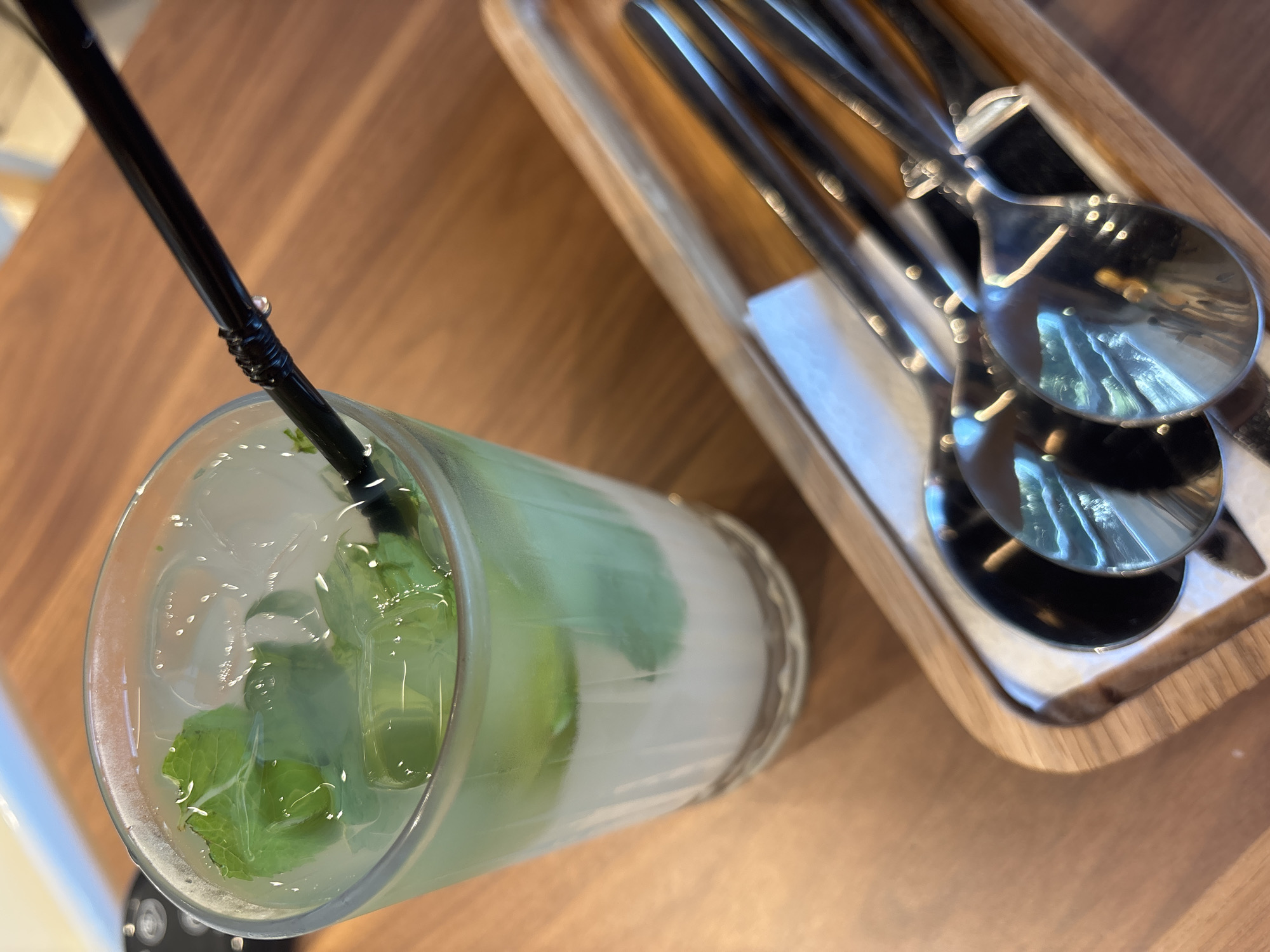 A close-up, overhead shot features a clear glass filled with a layered beverage, consisting of a white base and a pale green liquid with mint leaves and ice. A black straw pierces the top layer of the drink. The glass sits on a wooden table. To the right of the glass, a wooden tray holds several metallic spoons, their reflective surfaces showing distorted reflections of the surroundings. The background is a warm-toned wooden surface. No people are visible in the frame. The scene appears to be in a cafe or restaurant setting, possibly located in Naberezhnye Chelny, Russia.