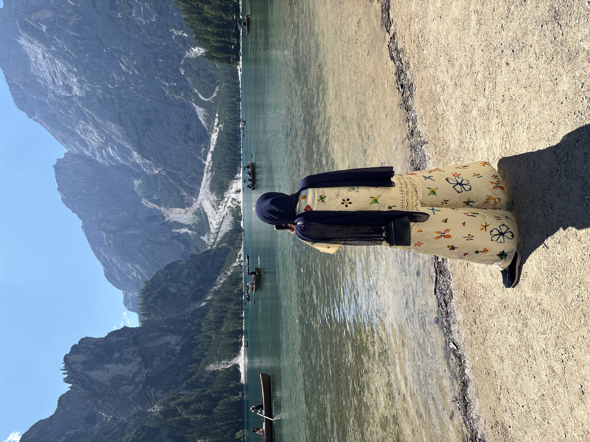 A person wearing a dark hijab, a light-colored loose-fitting outfit with colorful floral patterns, and dark shoes stands on a sandy shore facing away from the camera. They are holding a phone up to take a picture. In the background, a clear turquoise lake is dotted with several rowboats, some carrying passengers. The lake is surrounded by a dense forest of green trees. Towering, rugged mountains with steep rocky slopes and patches of snow dominate the horizon, under a bright blue sky with a few wisps of clouds. The scene appears to be a serene natural landscape. This location is likely Lago di Braies (Pragser Wildsee) in the Prags valley, South Tyrol, Italy, famous for its stunning mountain scenery and clear waters.