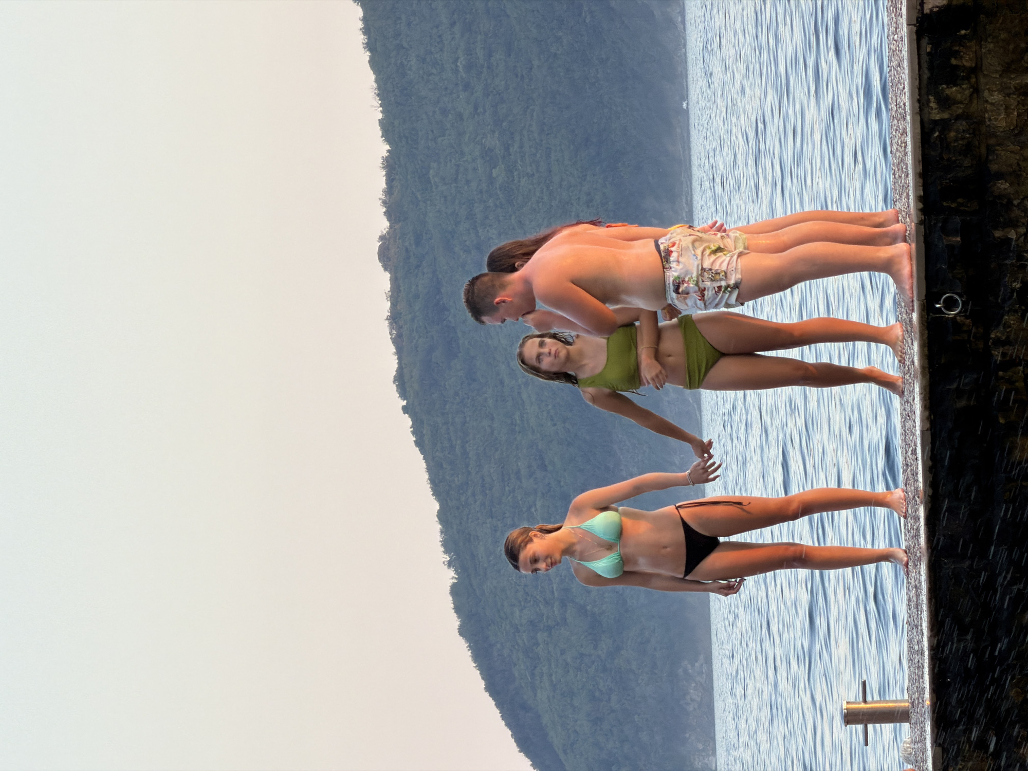 In Boreti, Montenegro, four individuals stand on a stone pier overlooking the Adriatic Sea at what appears to be sunset. On the left, a woman in a teal bikini and black bottoms holds hands with a woman in a lime green bikini. To their right, a man in patterned swim trunks stands facing them, and behind him, a woman in a pink swimsuit is partially visible. The sea is calm with gentle waves, and a lush, green hillside forms the background under a pale sky transitioning from light blue to a soft yellow. A small boat is visible in the distance on the water.