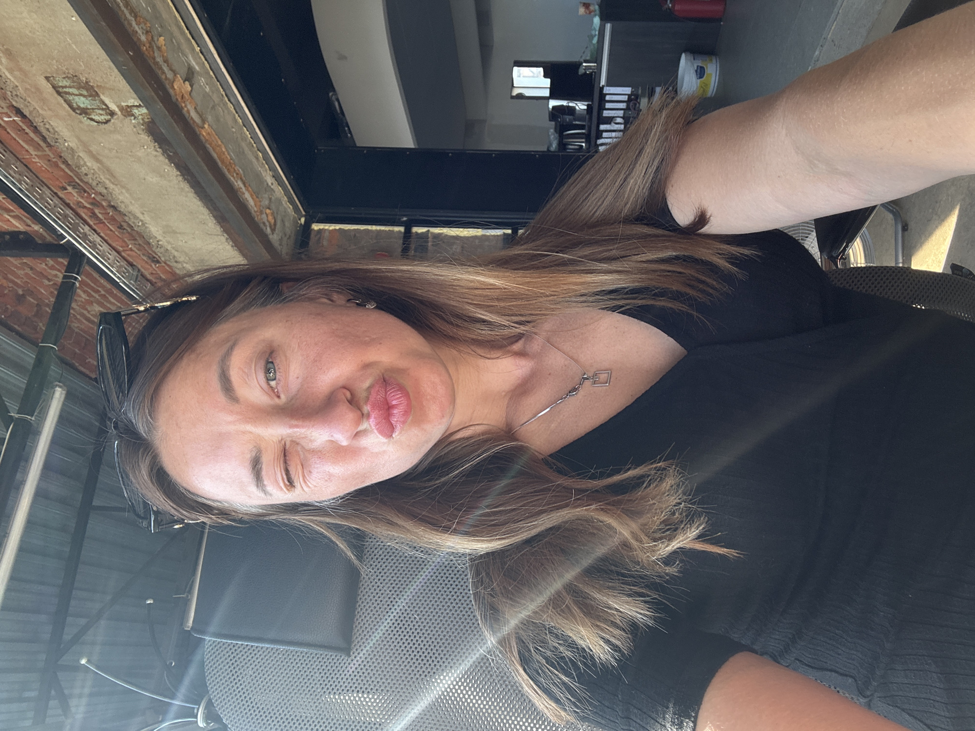 A selfie of a woman with light brown hair winking and puckering her lips. She is wearing a black top and a silver necklace with a square pendant. Sunglasses are perched on her head. The background shows a brick wall, a metal structure, and glimpses of an interior space. Based on these elements, it is difficult to identify the specific location in the city of Chaikovsky, Russia.