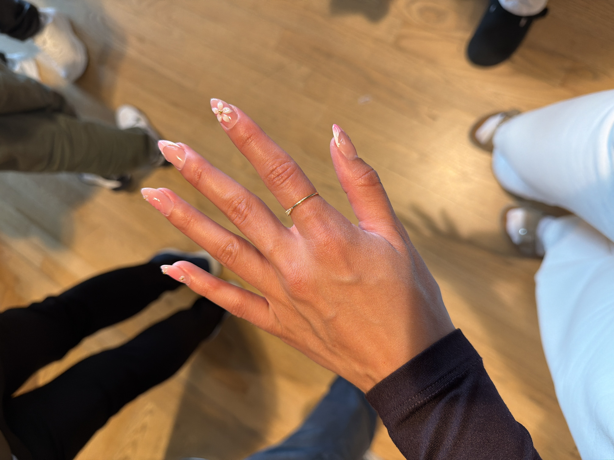 In Gardermoen, Norway, a person's hand with manicured nails featuring a delicate floral design is prominently displayed in the foreground. The hand is adorned with a thin gold ring. In the background, out of focus, are the legs of other individuals, one wearing black pants and the other green pants, along with their footwear. The setting appears to be indoors, with a light wooden floor.