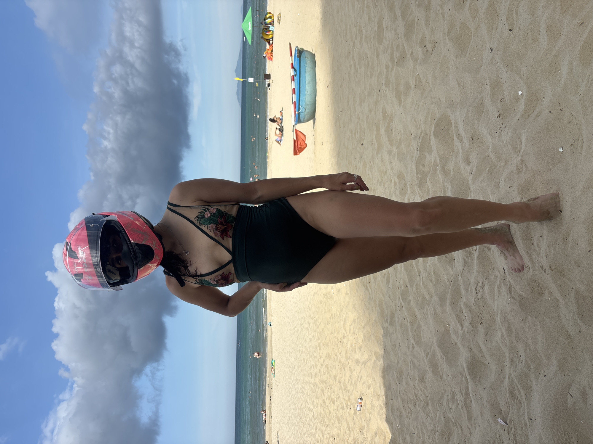 The image shows a woman in a black swimsuit and a pink helmet with a floral design. She is standing on a sandy beach with the ocean in the background. The sky is blue with some clouds. There are other people in the water and on the beach. There is also a blue inflatable boat on the sand. The scene appears to be a sunny day at a beach resort. The woman's tattoo is visible on her shoulder and back. The overall mood is relaxed and casual.