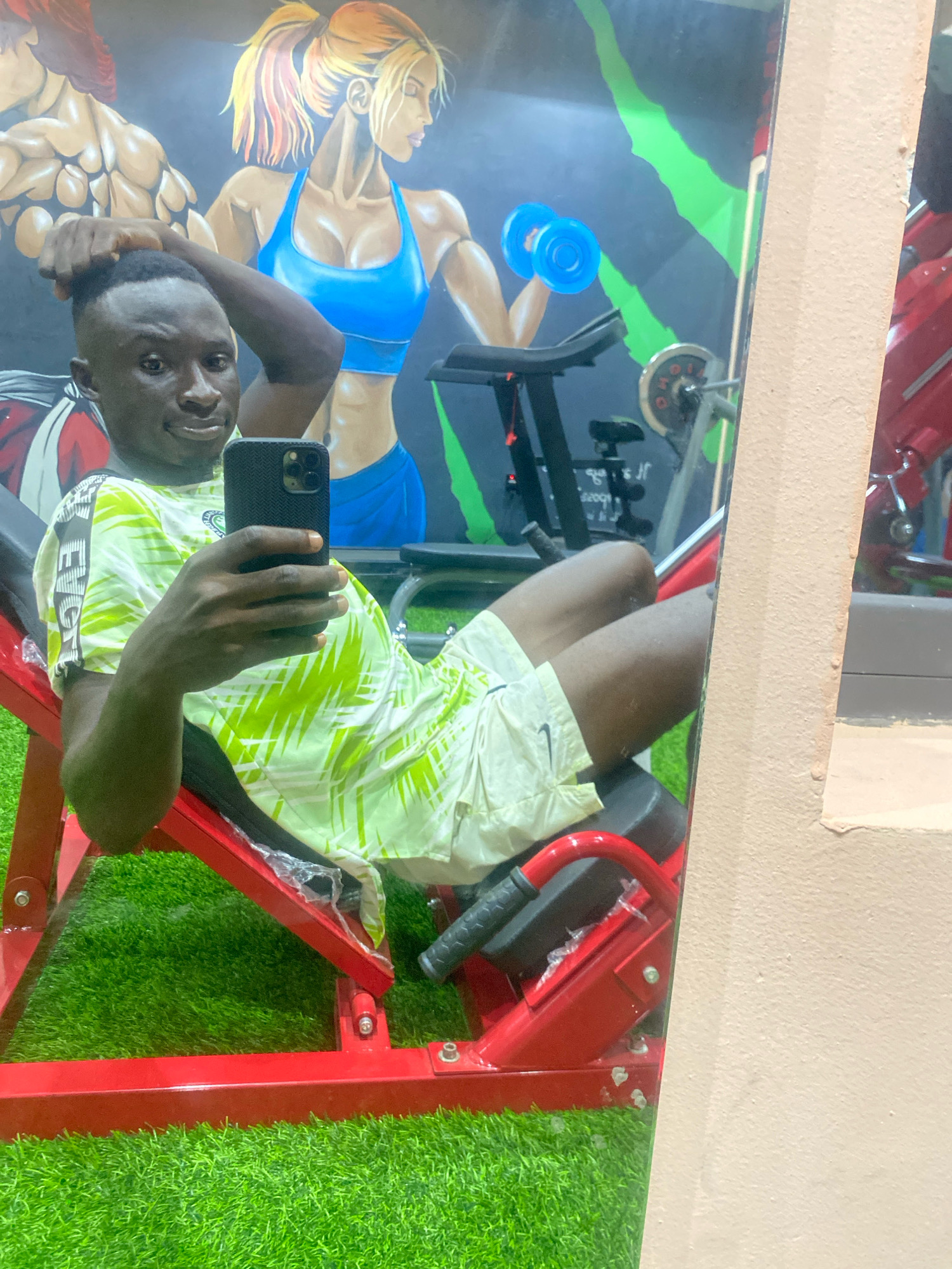 The image shows a young Black male taking a selfie in a gym. He is sitting on a red leg press machine, which is placed on artificial green turf. The man is wearing a light green and white patterned t-shirt and light-colored shorts. He is holding a black smartphone in his right hand, with his left hand resting on his head. Behind him is a mural depicting muscular figures, one male and one female, with a blue dumbbell and green abstract shapes. To the right of the man, part of a pinkish wall is visible. The gym appears to be located in the city of Barden Yana, Nigeria.