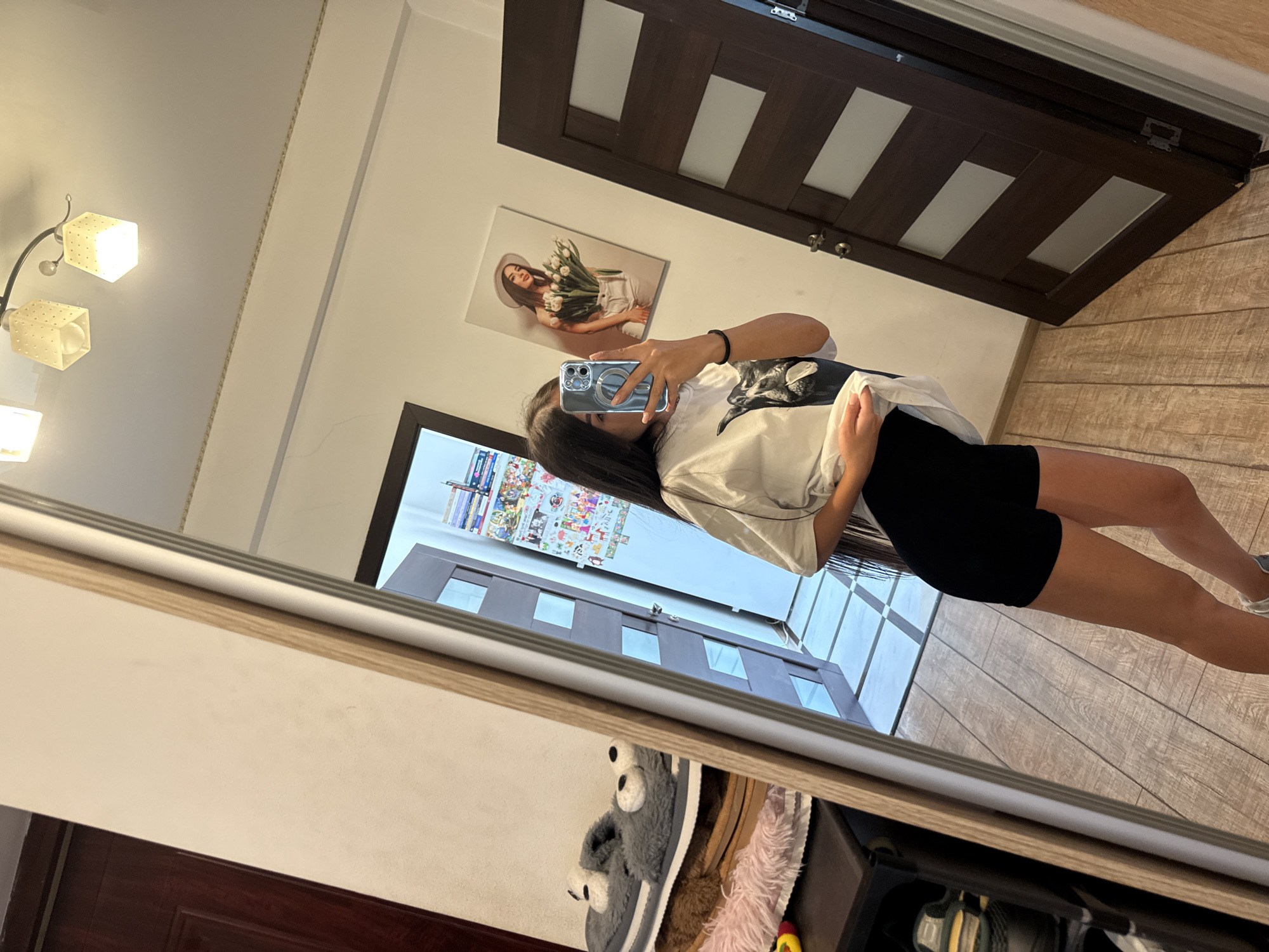 A young woman with long dark hair is standing in front of a full-length mirror, taking a selfie with her phone. She is wearing a white t-shirt with a graphic print and black shorts. The mirror reflects her and the room behind her. To the left of the mirror, there is a shelf with slippers and other items. In the background, there are two doors, one a dark wood with frosted glass panels, and the other a lighter colored door with multiple decorative elements. A framed picture of a woman holding flowers hangs on the wall. The floor is tiled in a checkered pattern in one area and wooden planks in another. The lighting in the room appears to be from overhead fixtures. The overall setting suggests an indoor residential space, possibly a bedroom or hallway. No specific identifiable landmarks or elements in the image definitively place the location in Chișinău, Moldova.