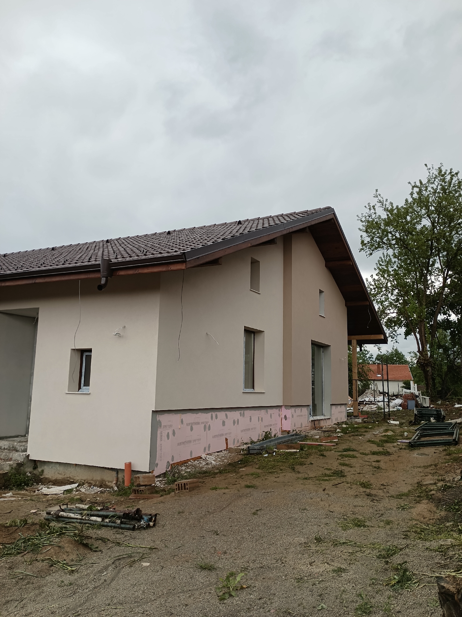 The image depicts a single-story house under construction in Mihaylovtsi, Bulgaria. The house has a brown tiled roof, tan exterior walls, and brown trim. The base of the house is lined with pink insulation material. Several windows and a sliding glass door are visible. Construction debris and materials are scattered around the house, including pipes, bricks, and metal framing. In the background, a tree and another house with a red roof can be seen. The sky is overcast. The ground is dirt and gravel.