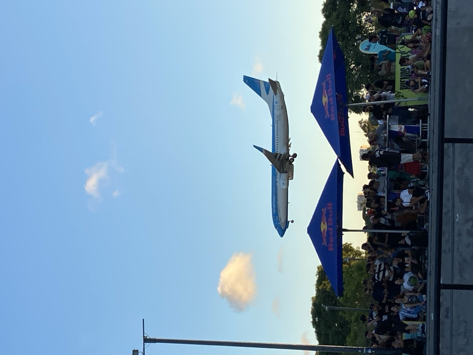 A passenger airplane with blue and white paint does an acrobatic maneuver in the sky near a large crowd of onlookers. The plane is upside down, its landing gear exposed. Two blue Red Bull banners are positioned near the spectators, suggesting the event is sponsored. Trees and buildings are visible in the background. The sky is blue with some clouds. A light pole is visible in the foreground.