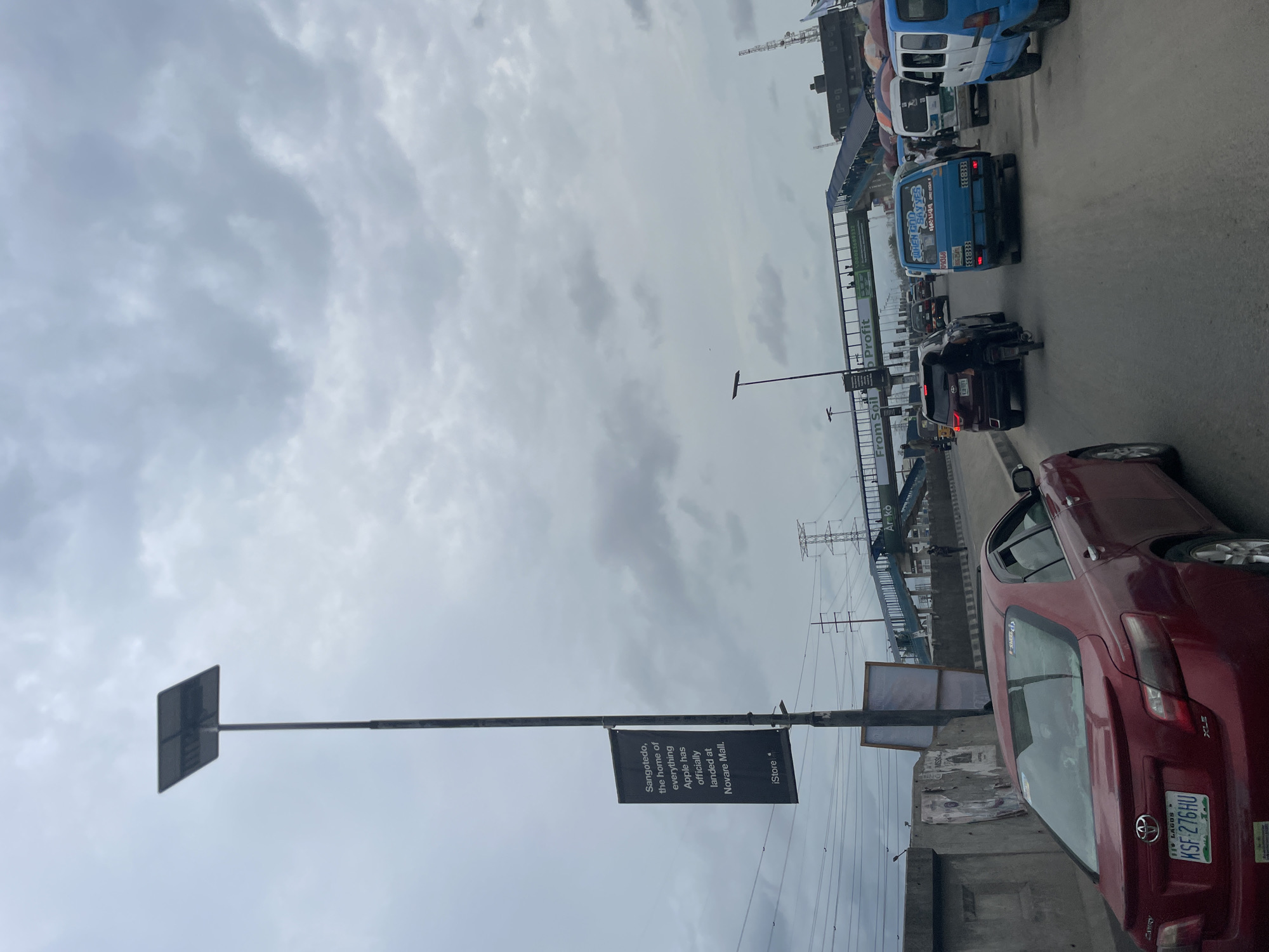 The image depicts a congested street scene in Lekki, Nigeria, specifically near Sangotedo. A red Toyota Camry is parked or slowly moving in traffic, its license plate reading "KSF-276HU." Adjacent to the road, an advertising banner promotes the opening of an Apple iStore at Novare Mall in Sangotedo. In the background, one can see a bridge infrastructure, power lines, and numerous vehicles, including buses and cars, all contributing to the heavy traffic. The sky is overcast with cloudy conditions. The presence of signage mentioning "From Soil" and "Profit" further characterizes the roadside environment.