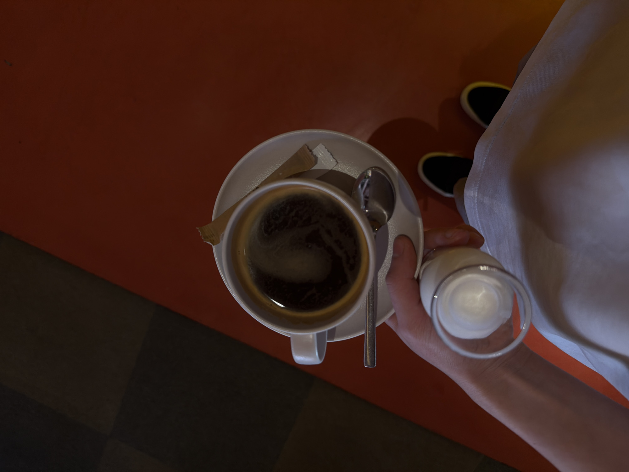 An overhead shot shows a cup of black coffee on a white saucer, with a silver spoon, and packets of sugar. A hand holds a small glass of milk near the coffee cup. The surface beneath is primarily orange, transitioning to a black and gray checkered pattern. In the background, parts of a person are visible, including black shoes with white trim and a white garment. The location is Yerevan, Armenia.