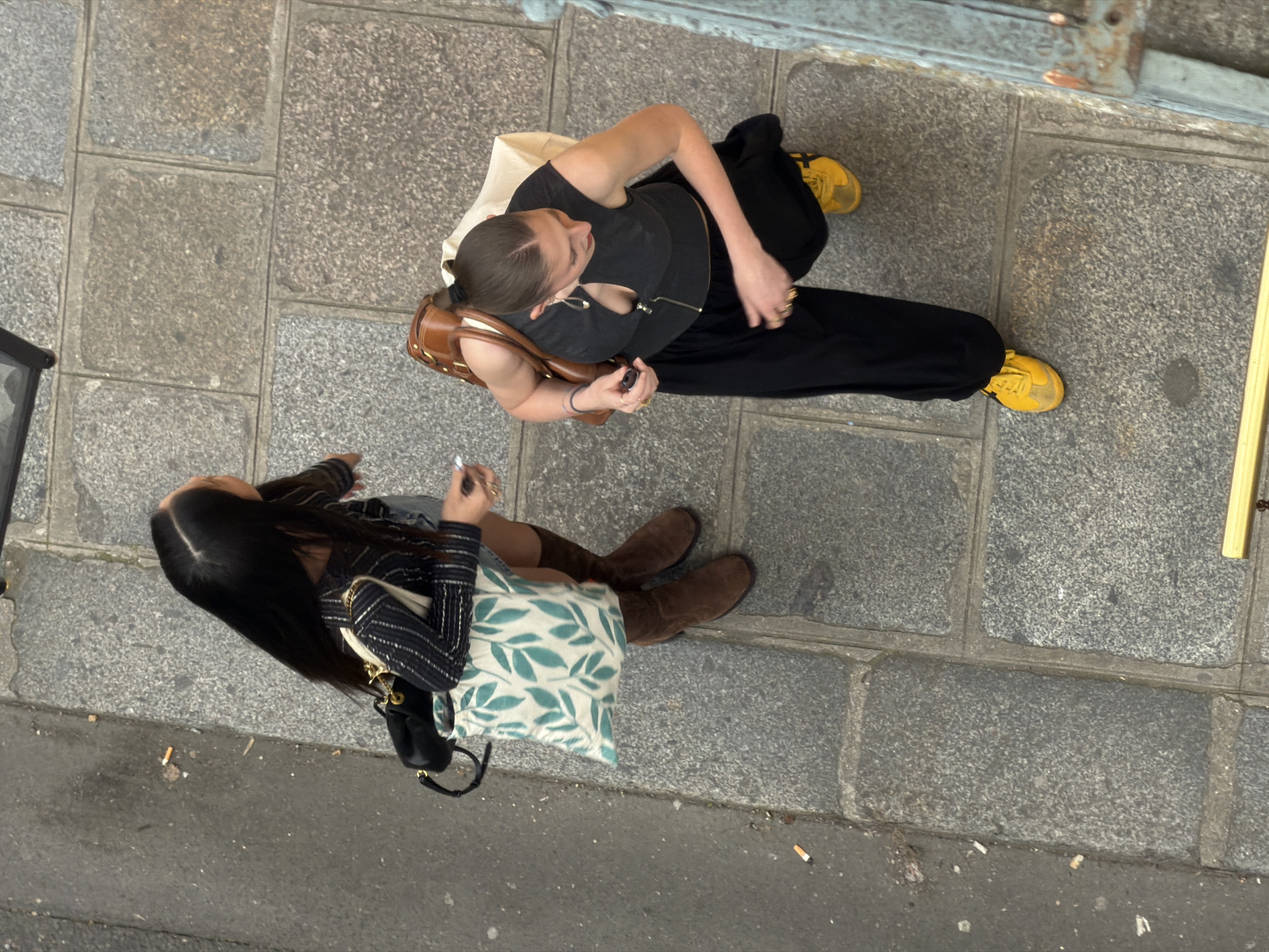 The image is an aerial view of two women on a stone-paved street in what appears to be Paris, France. The street is made of uneven grey stone pavers. Litter, including cigarette butts, is scattered on the side of the street. One woman wears black clothing and yellow sneakers. She has a brown bag and a tan colored shoulder bag. The other woman wears a striped top, skirt, and brown boots and has a patterned tote bag. Both women are looking at their phones.