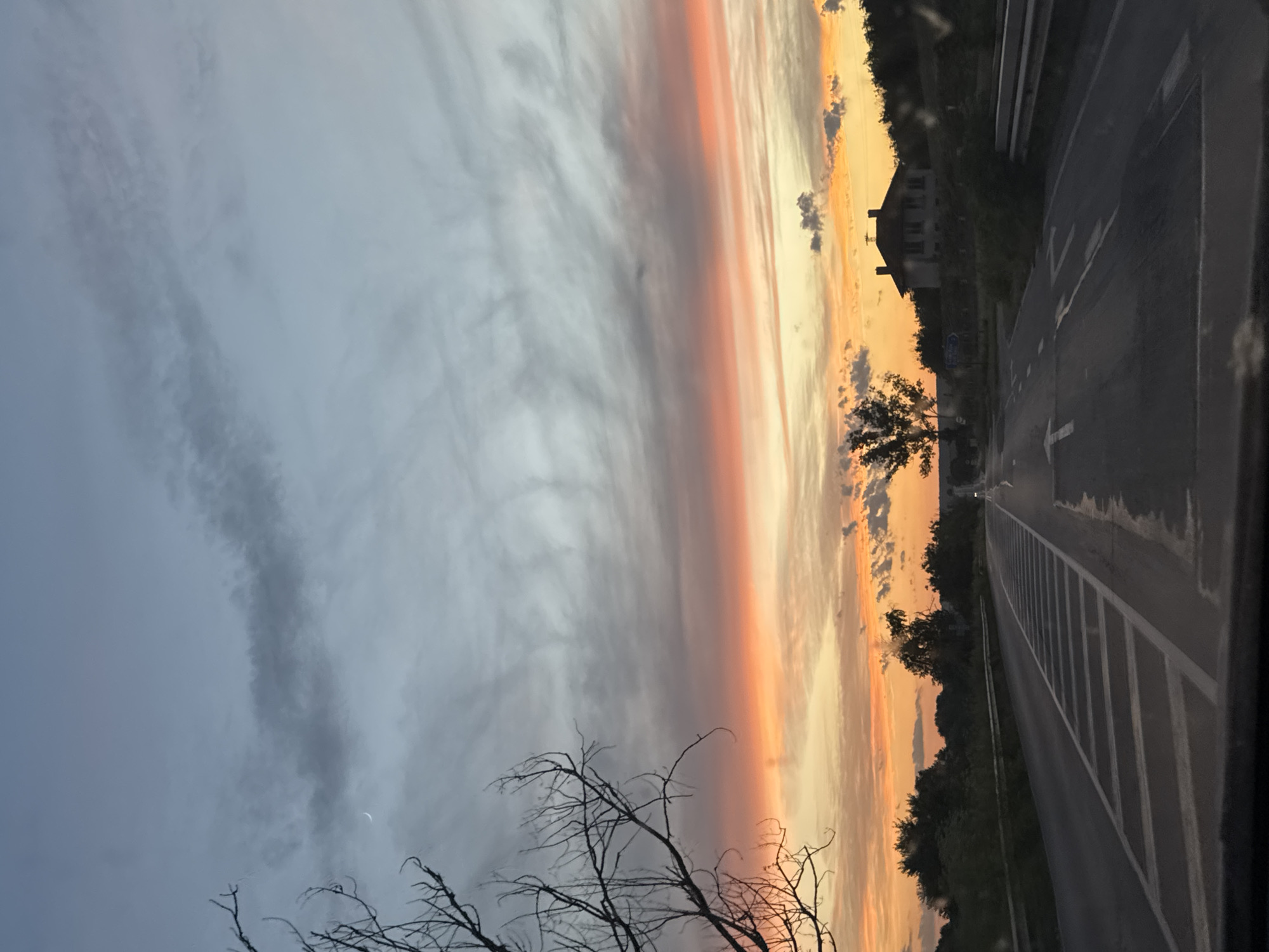 The image depicts a scenic view of a road in the city of Beli Lom, Bulgaria, during sunset. The sky is a gradient of colors, transitioning from a pale blue at the top to vibrant orange and pink hues near the horizon, indicating the sun is setting. A thin crescent moon is visible in the upper left portion of the sky. Wispy clouds stretch across the sky, adding texture and depth.
In the foreground, a two-lane road stretches into the distance, with white lane markings clearly visible. The road appears to be wet in places, suggesting recent rain. On the left side of the road, a bare tree with intricate branches stands out against the colorful sky. On the right side of the road, a guardrail runs alongside, and further in the distance, a two-story house with a dark roof is situated on a gentle slope, surrounded by lush greenery and trees. The overall atmosphere is tranquil and picturesque, capturing the beauty of a rural sunset in Bulgaria. There are no people or animals visible in the image.