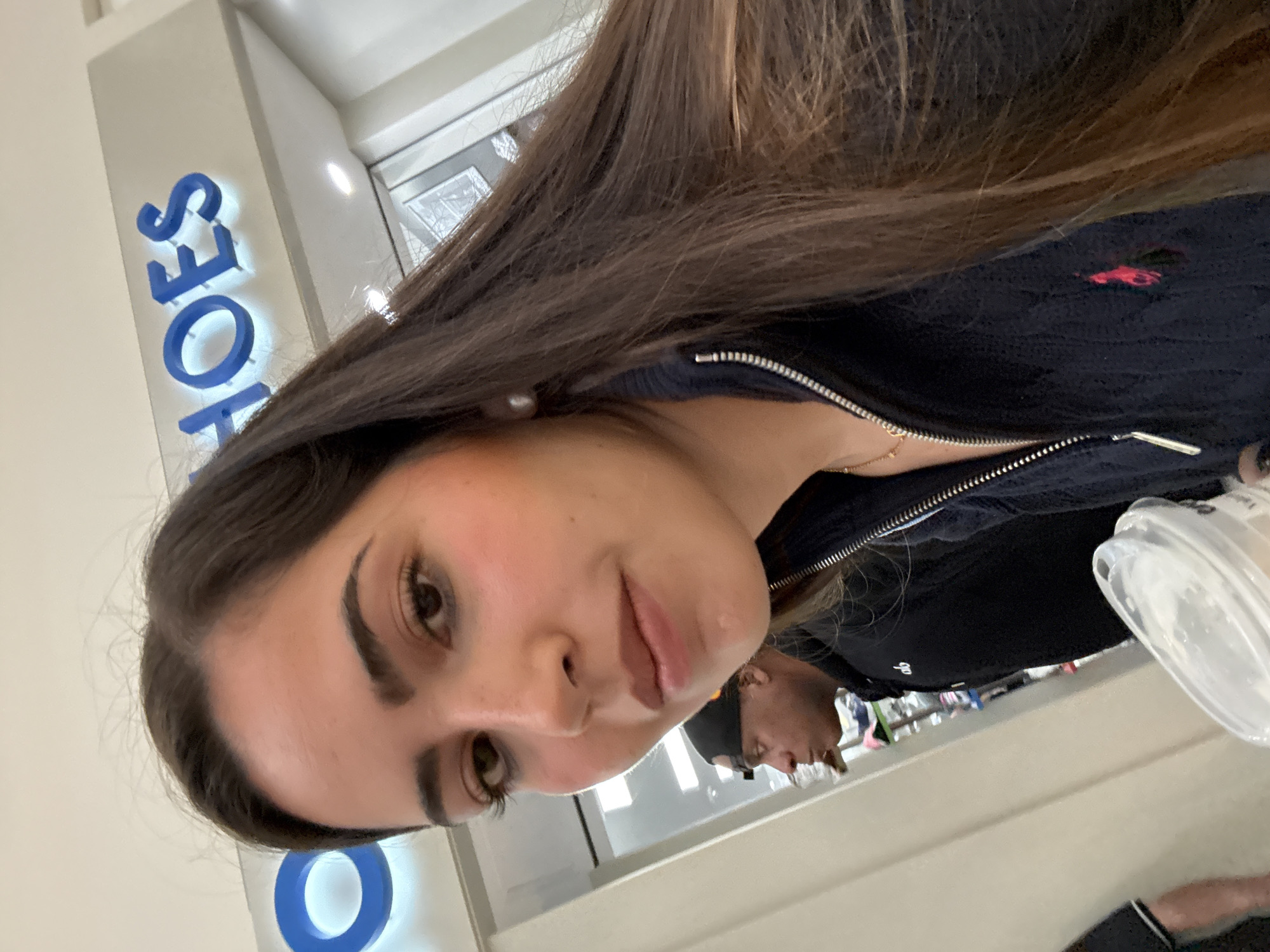 The photograph captures a selfie-style view of a person with brown hair, light skin, and notable eyebrows. She is wearing a dark-colored, possibly navy blue, zip-up jacket, identifiable by a small red logo on the left side and a gold necklace. The setting appears to be within a retail environment, specifically a "SHOES" store. In the background, partially obscured, is another individual wearing a black beanie and a black t-shirt. The photo's angle and the presence of the retail store sign suggest the location could be a shopping mall. The picture was taken in San Antonio, United States of America. A clear plastic container is visible in the corner of the photograph.